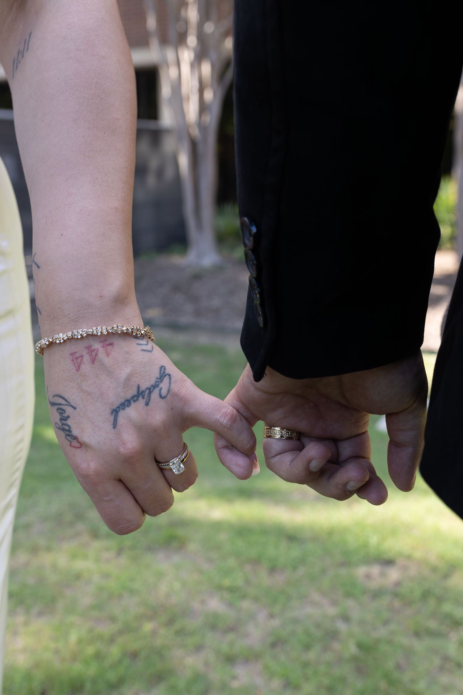 Couple holding hands with tattoos and wedding rings visible, standing on green grass outdoors.