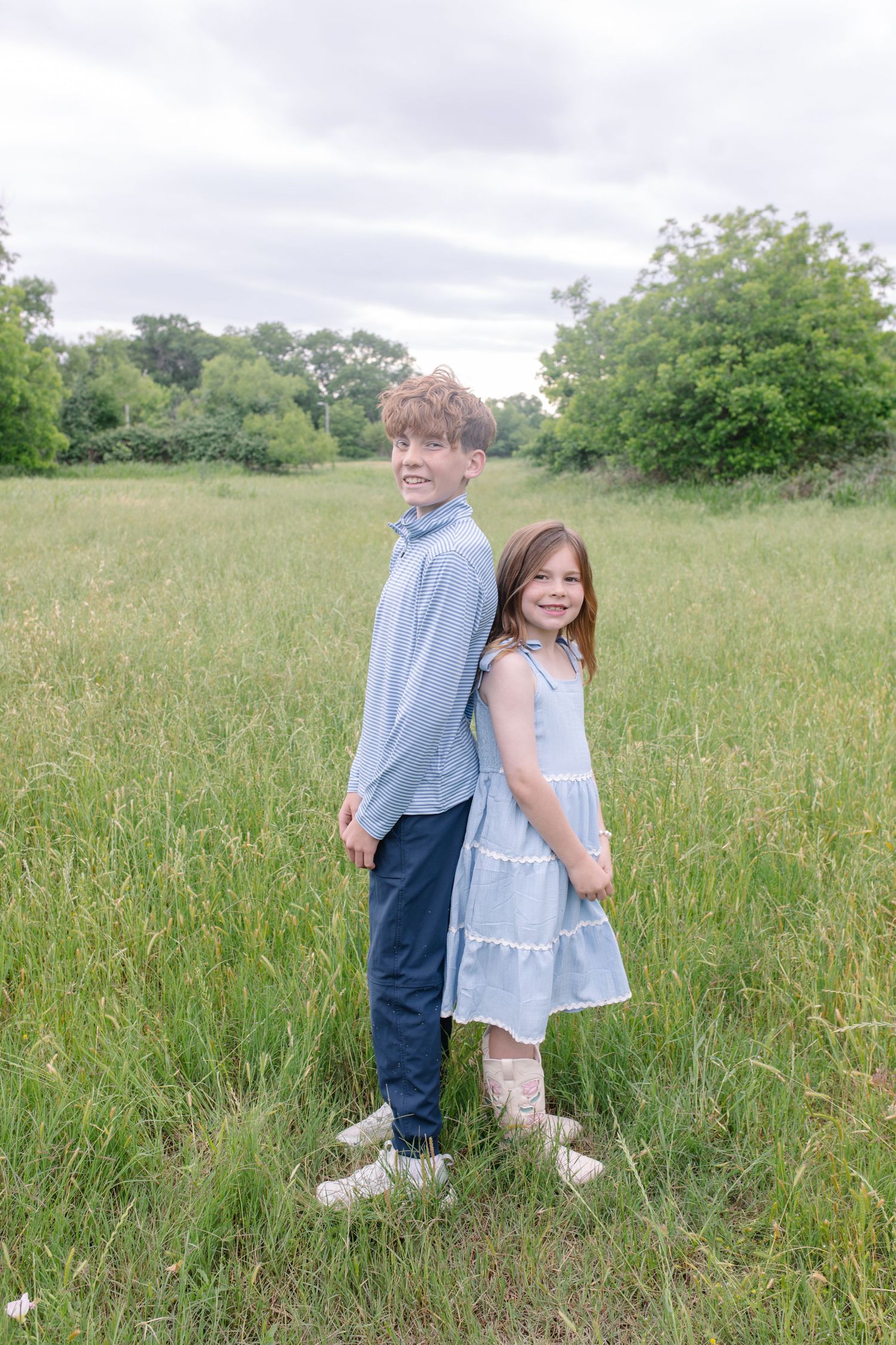 Two children in blue outfits stand back-to-back smiling in a lush green meadow on a cloudy day.