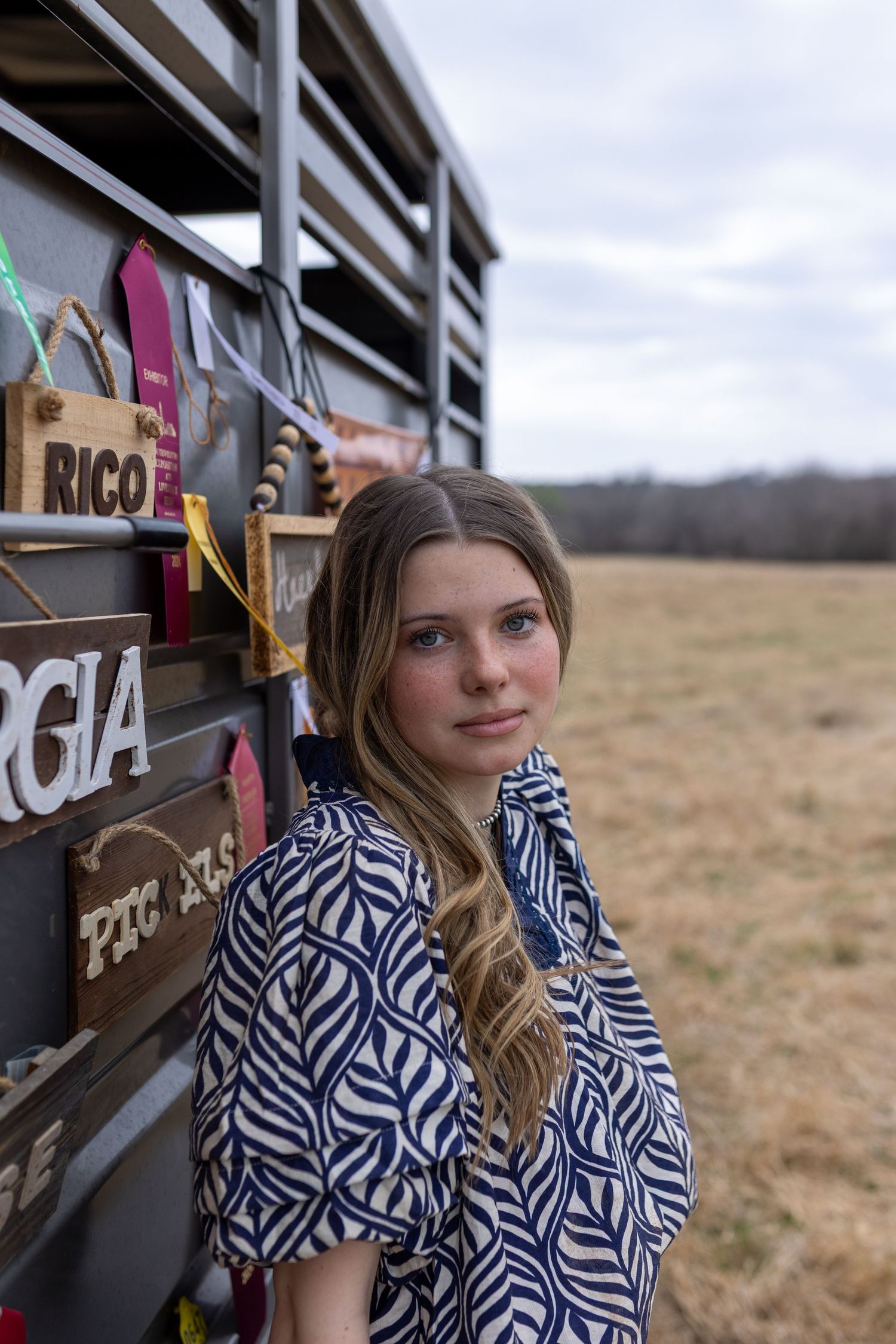 A person in a blue and white patterned top poses near a brown horse trailer in a rural field setting.