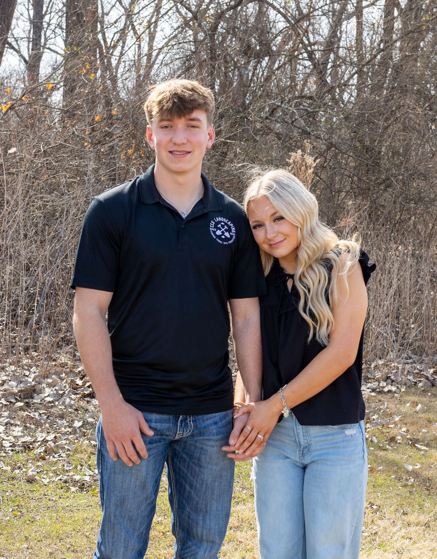 Family photo session outdoors with people wearing coordinated black shirts and jeans against a wooded backdrop.
