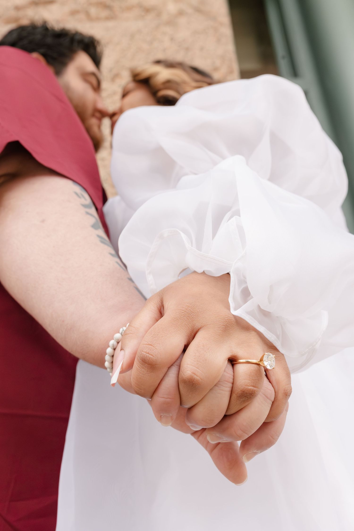 Close-up of newlyweds holding hands, bride in white dress with gold ring, groom in red suit with pearl bracelet.