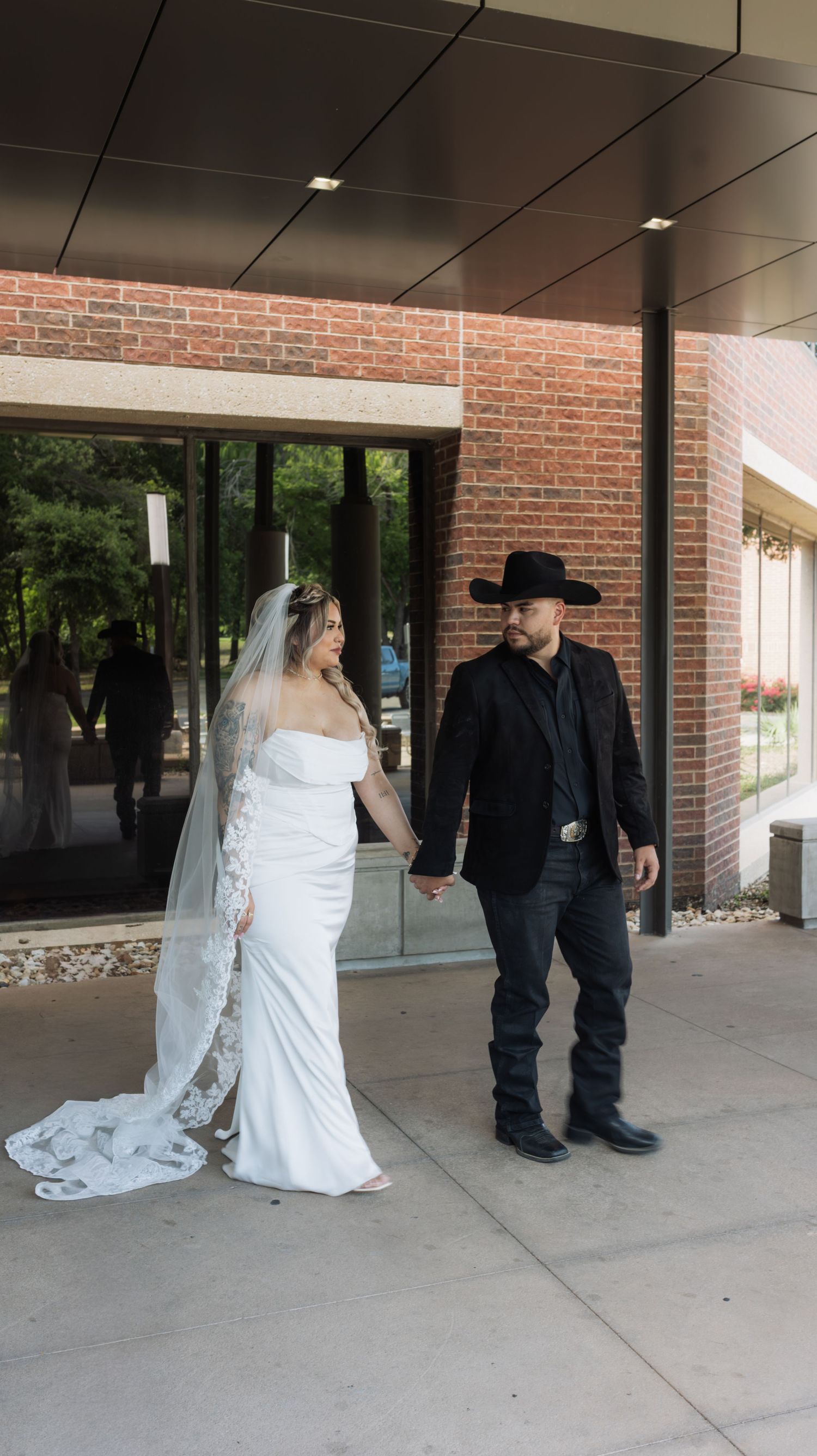 Bride in white gown with veil and groom in black suit and cowboy hat holding hands outside brick building.