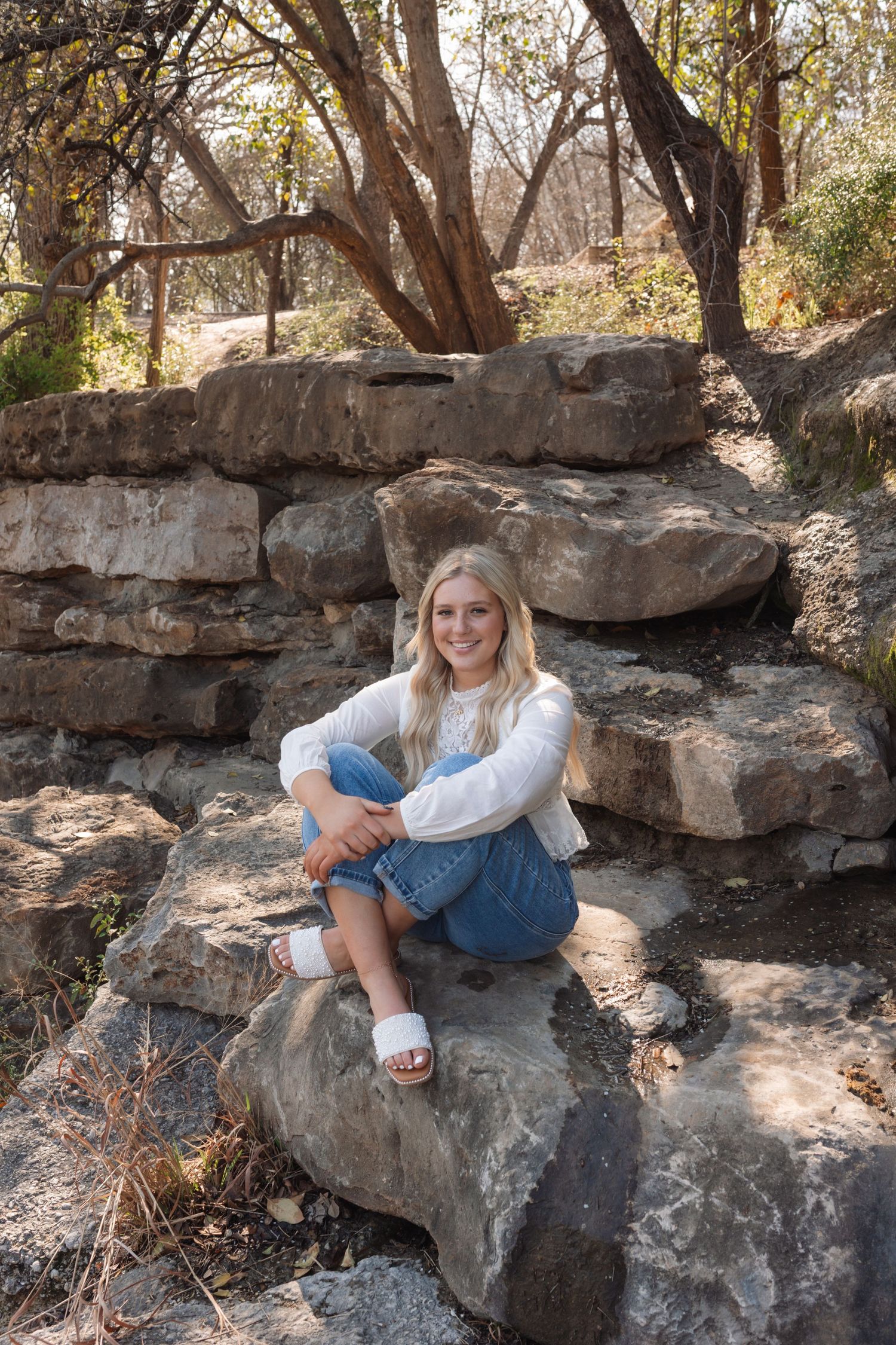 A person in jeans and white sweater poses casually on large rock formations in a wooded outdoor setting.