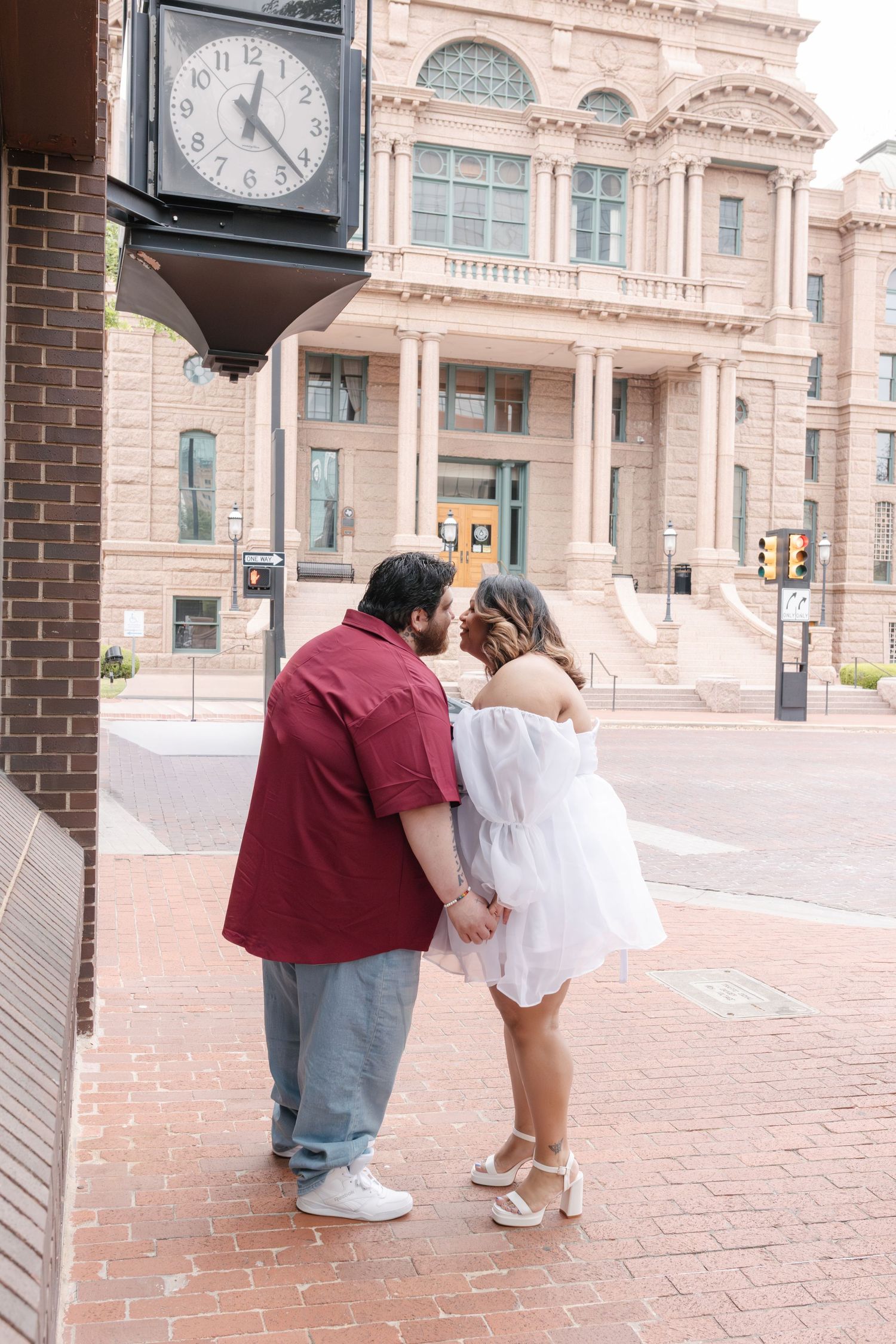 A couple shares an intimate moment on brick steps outside a historic courthouse building with large clocks.