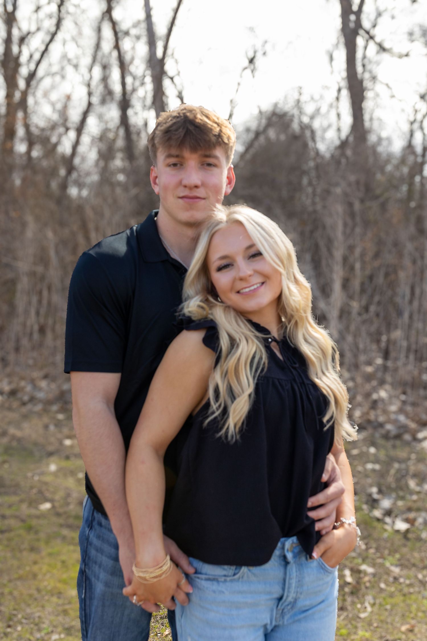 A couple embraces while posing together outdoors with trees and natural scenery in the background.
