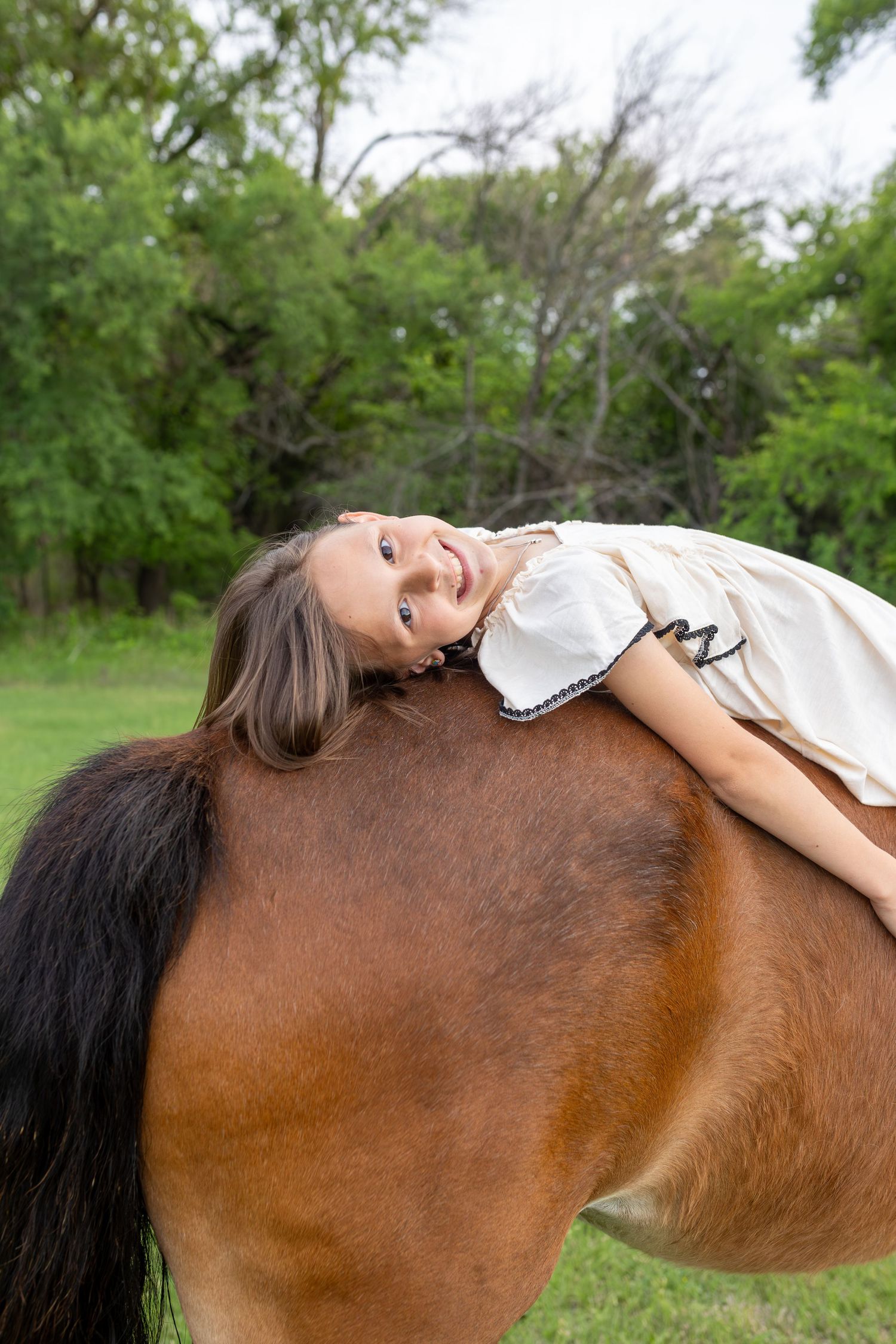 Young girl in white dress lying on back of brown horse, smiling outdoors with green trees background.