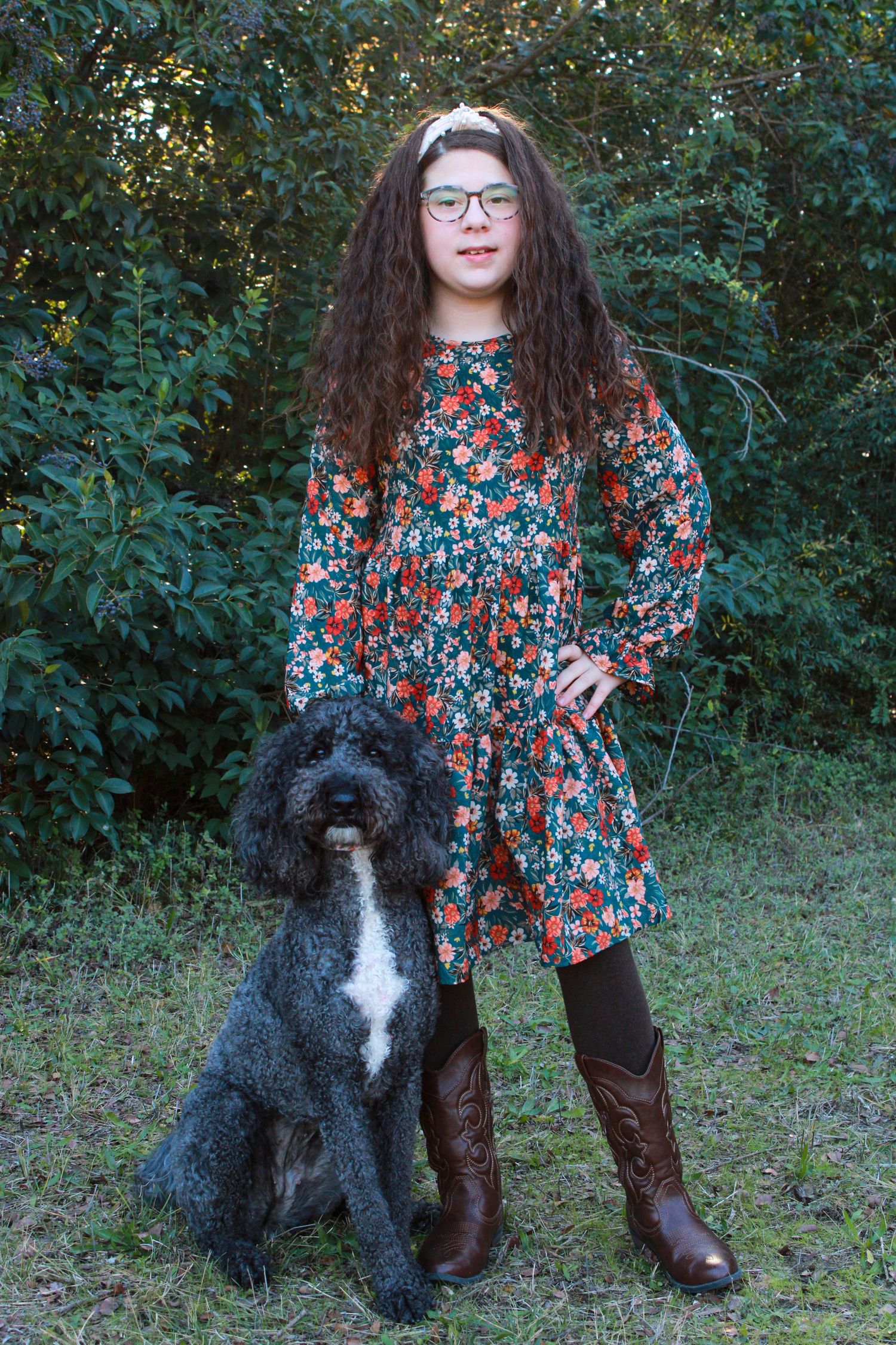 A black and white Bernedoodle dog poses outdoors with someone wearing a floral dress and brown boots.