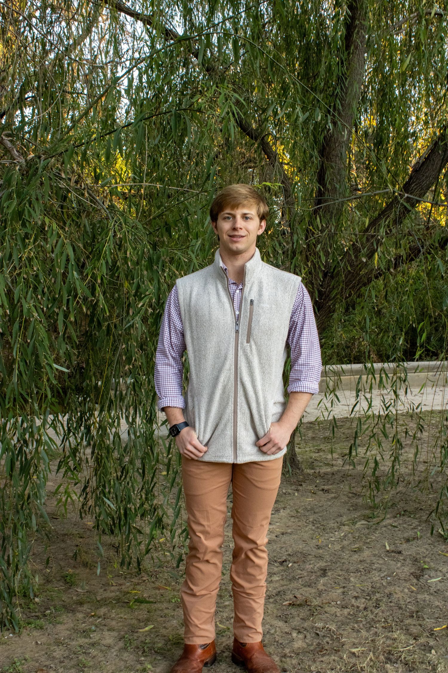 Someone wearing a light gray vest and blue button-down shirt stands in front of a weeping willow tree.