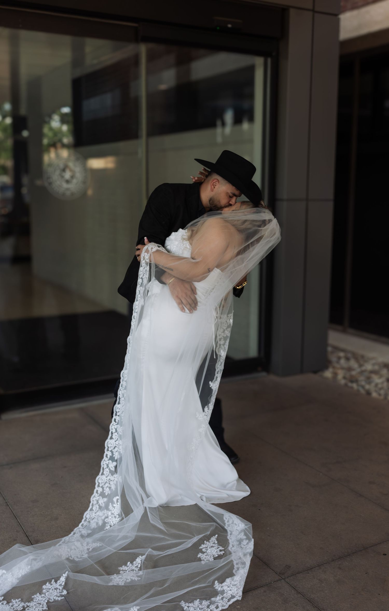 Bride in white lace gown and veil embraced by groom in black suit and hat outside a modern building.