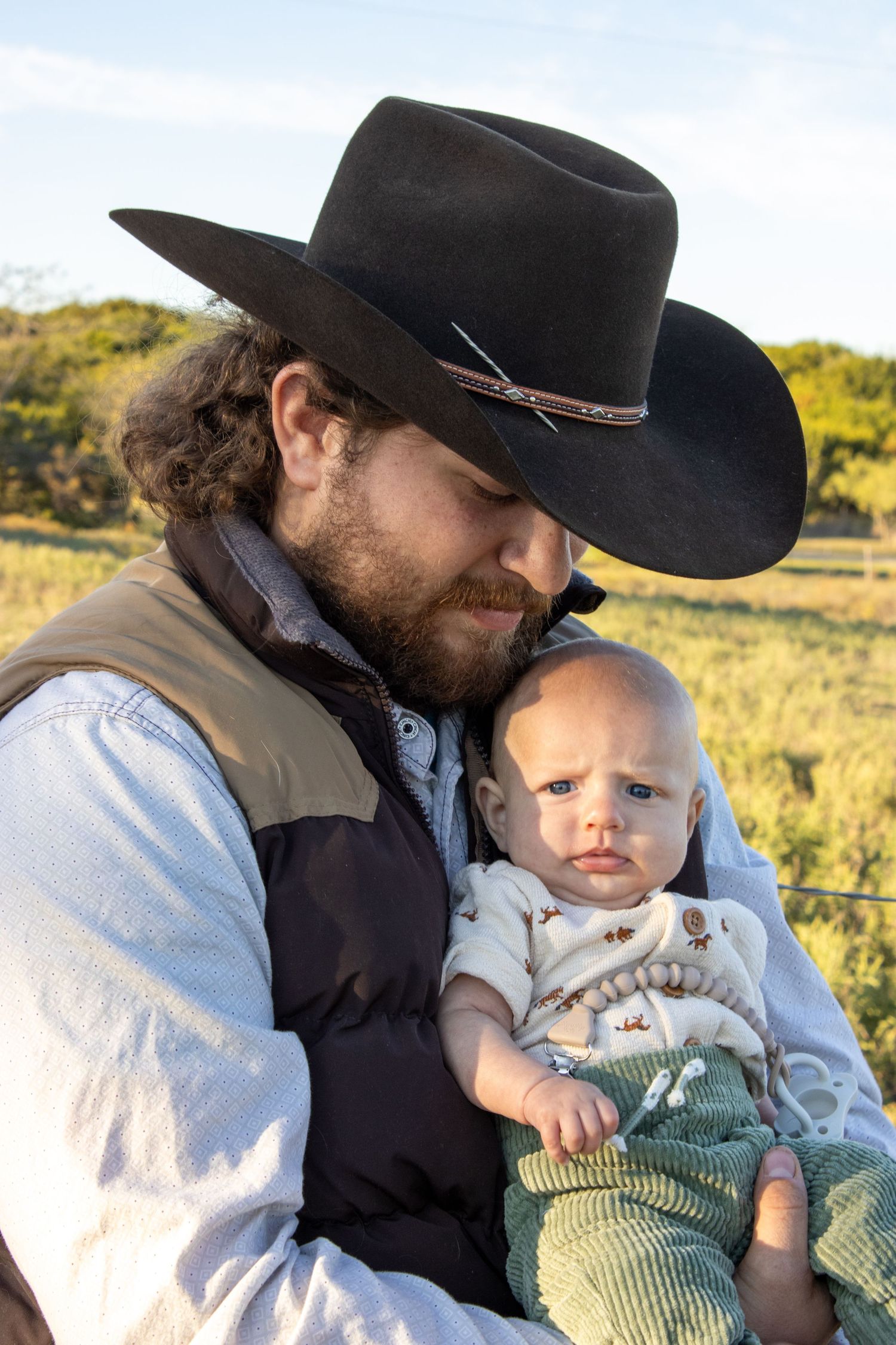 A tender moment captured in the sunlight showing affection between a cowboy-hatted figure and a baby outdoors.
