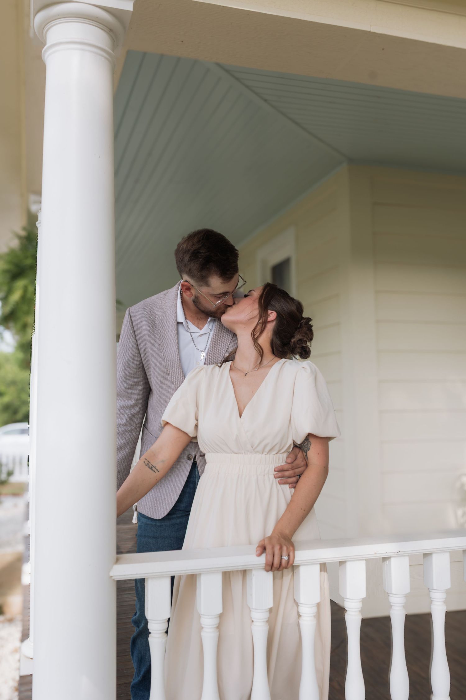 Couple sharing a romantic kiss on a white porch railing, man in gray blazer and woman in cream dress.