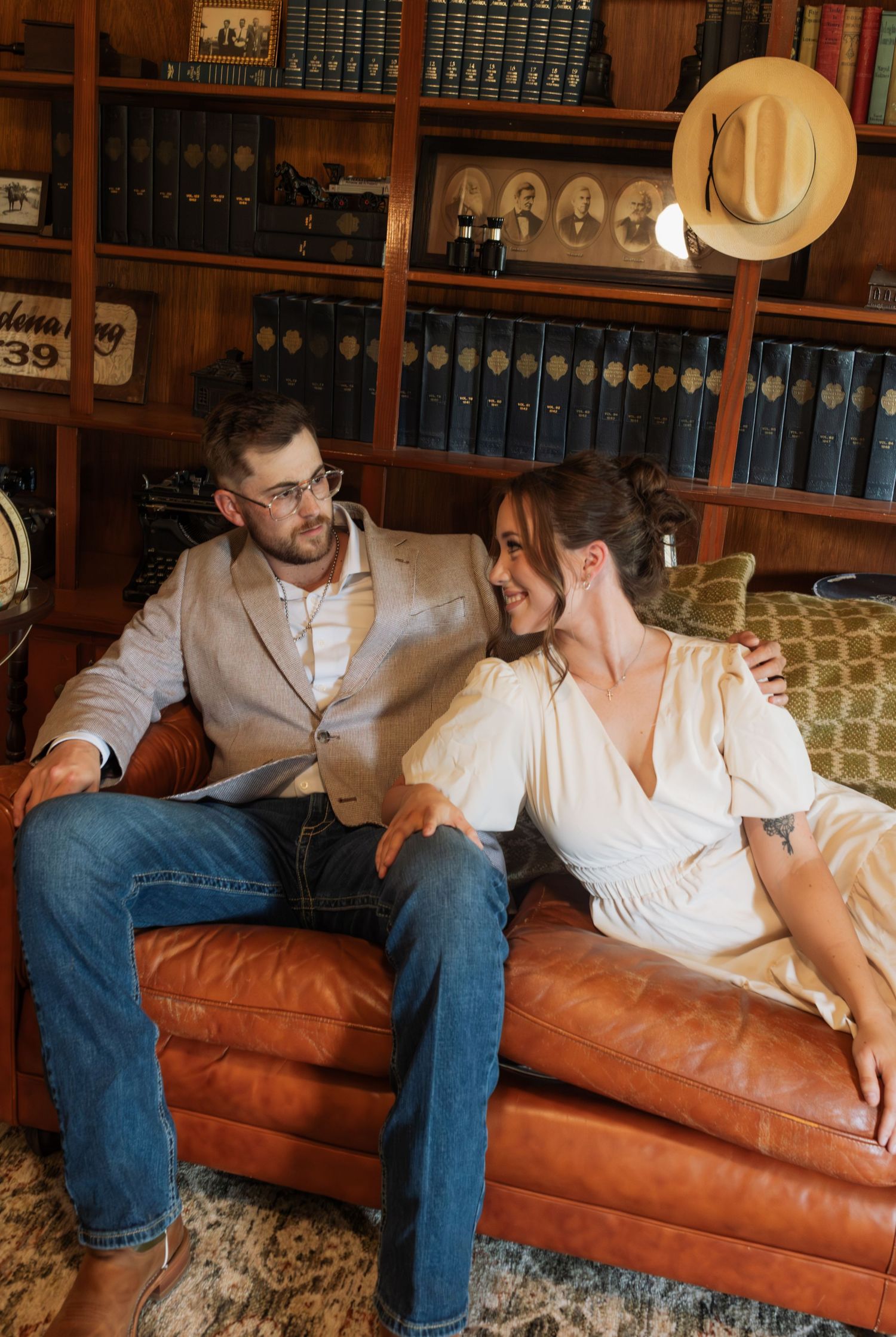 Couple sitting on leather couch in cozy library, man in blazer and jeans, woman in white dress, hat flying in air.