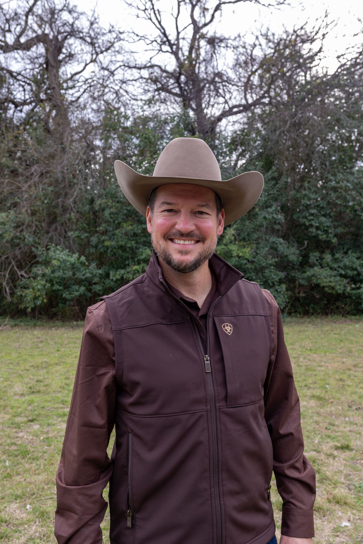 A person wearing a brown jacket and cowboy hat stands outdoors against a backdrop of trees and bushes.