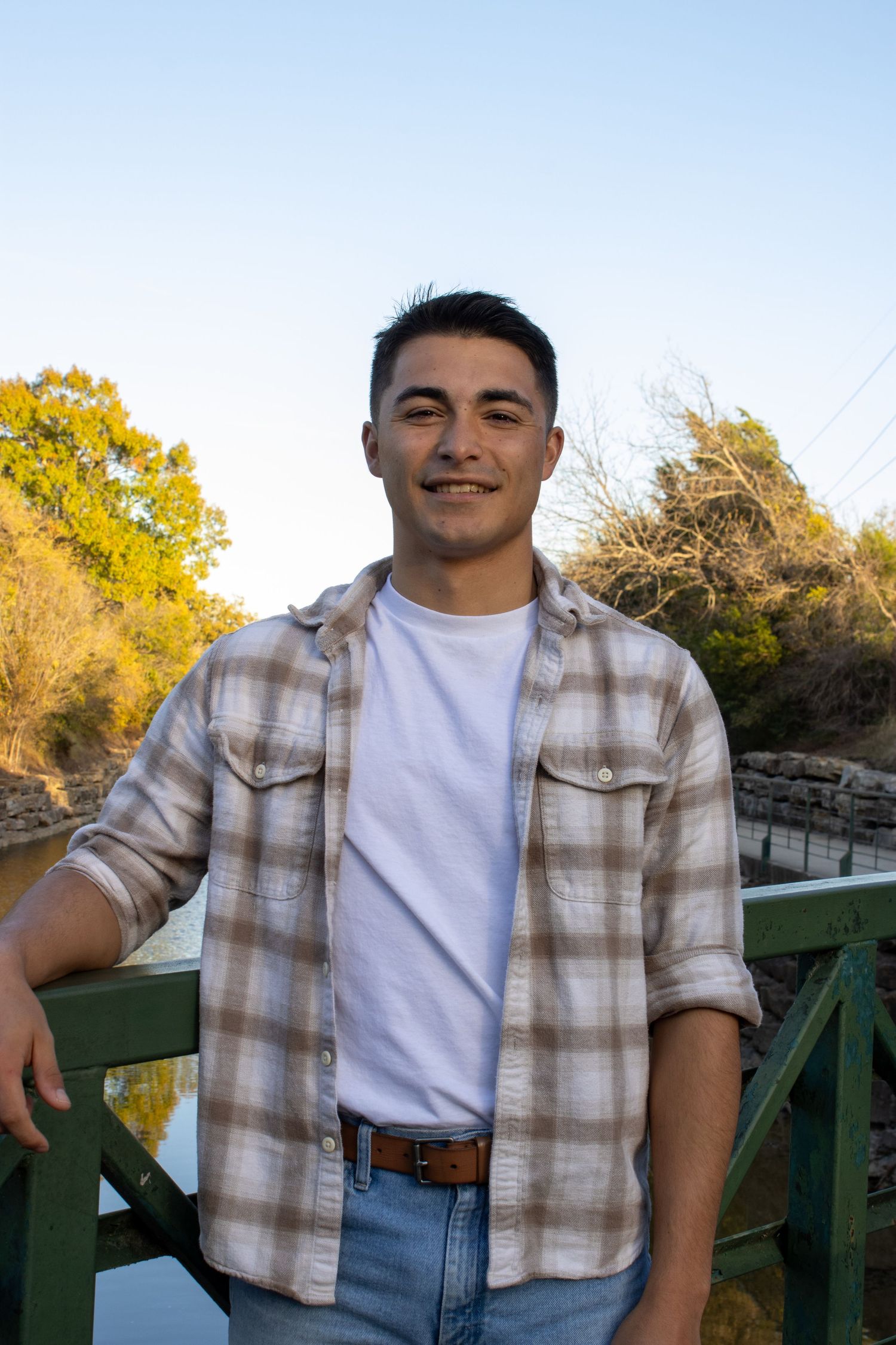 A person in a plaid button-up shirt and white t-shirt poses outdoors against a wooden railing on a sunny autumn day.