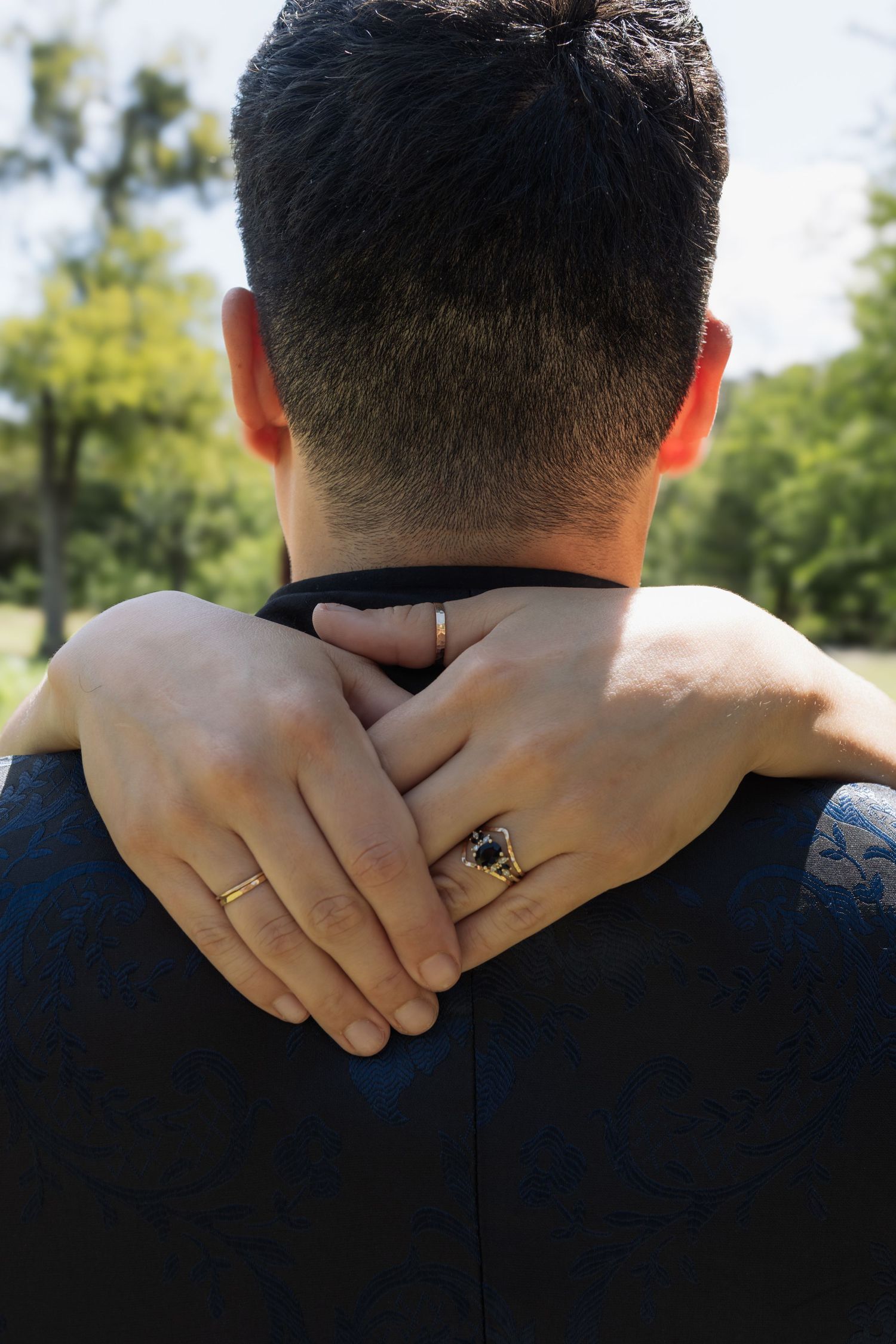 Couple embracing from behind, showing gold and black gemstone rings on clasped hands over man's suited shoulder.