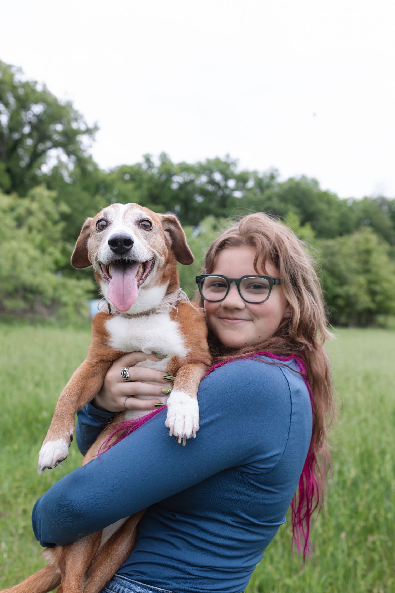 A smiling girl with glasses holds a happy brown and white dog outdoors in a lush green field.