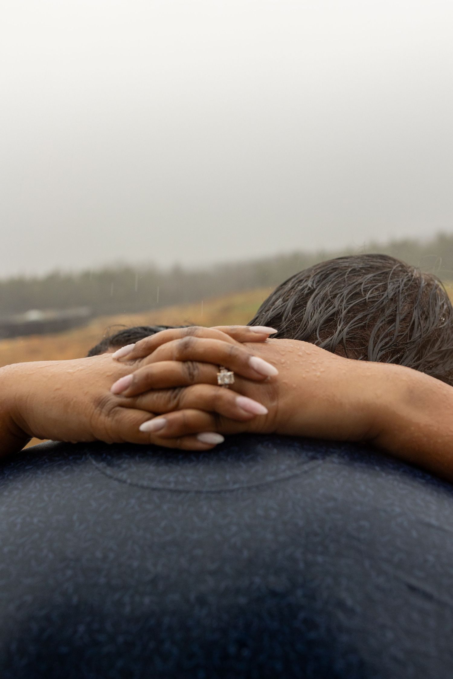 Hands gently rest on a person's shoulder wearing a dark blue shirt against a blurred background.