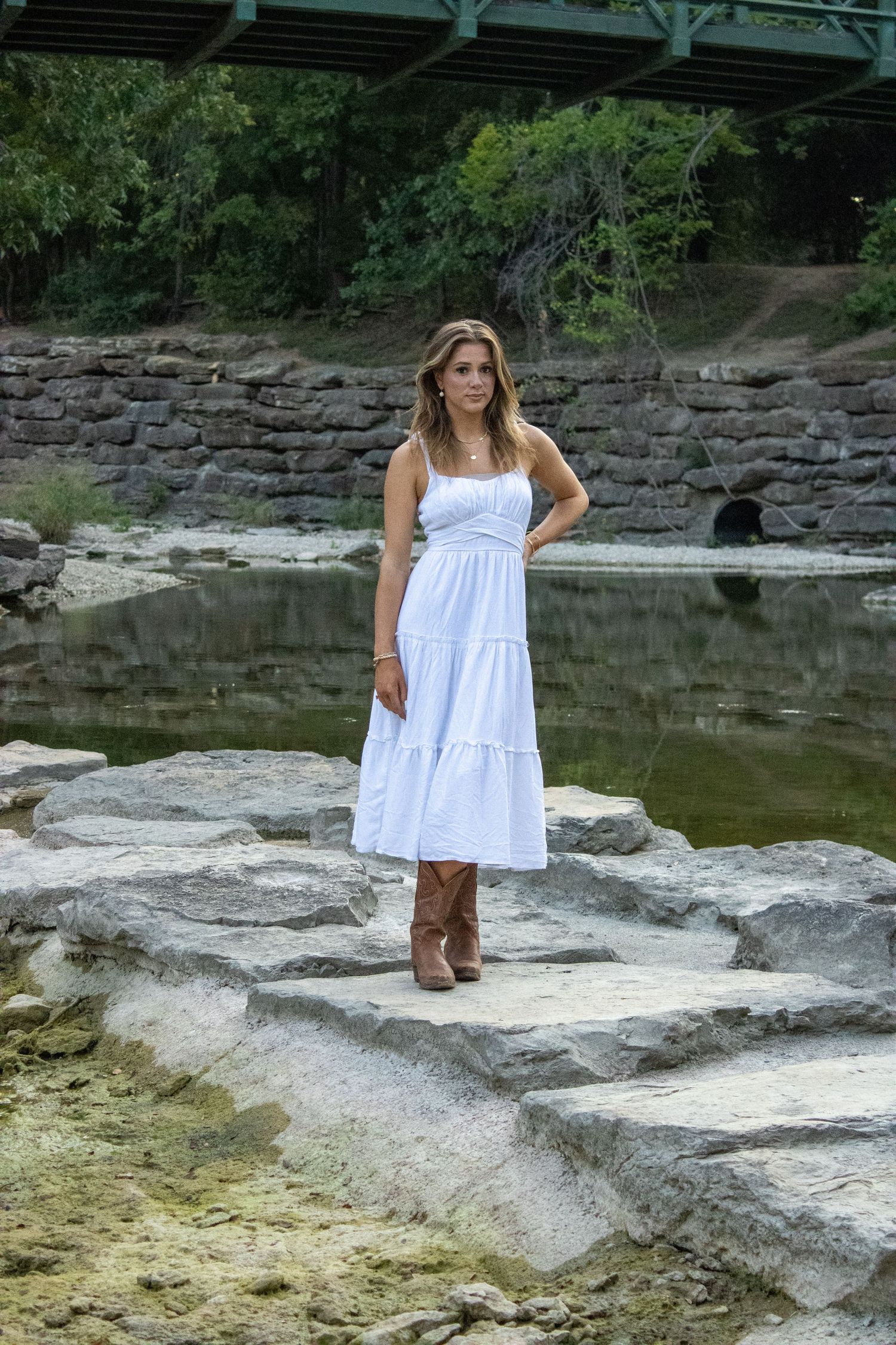 A scenic portrait by a stone wall and pond under a bridge.