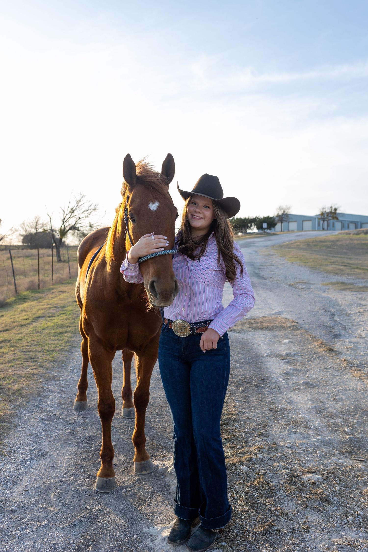 A brown horse stands with a cowgirl in western attire on a gravel path at sunset.