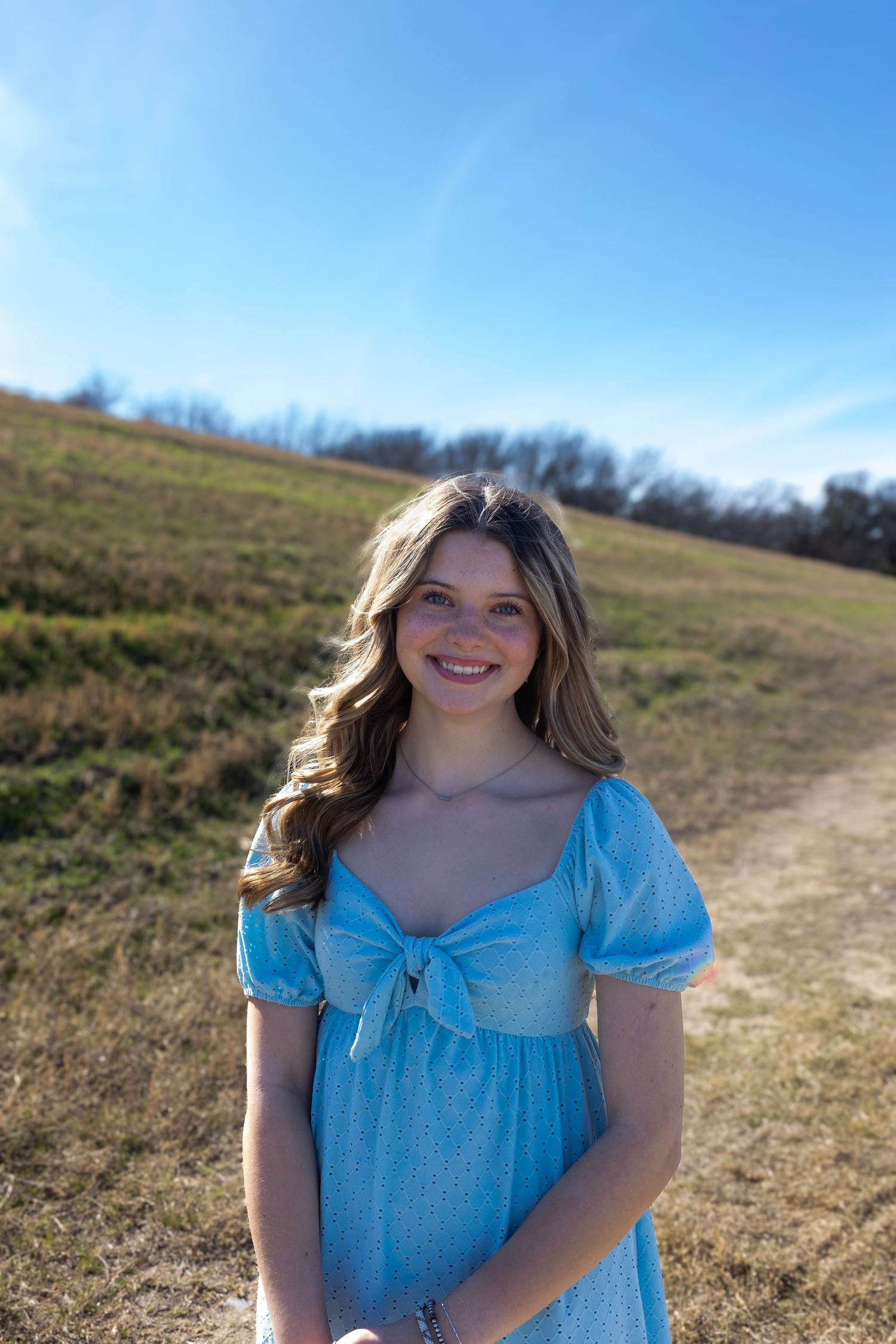 A person in a light blue dress with puff sleeves stands in a grassy field on a sunny day.
