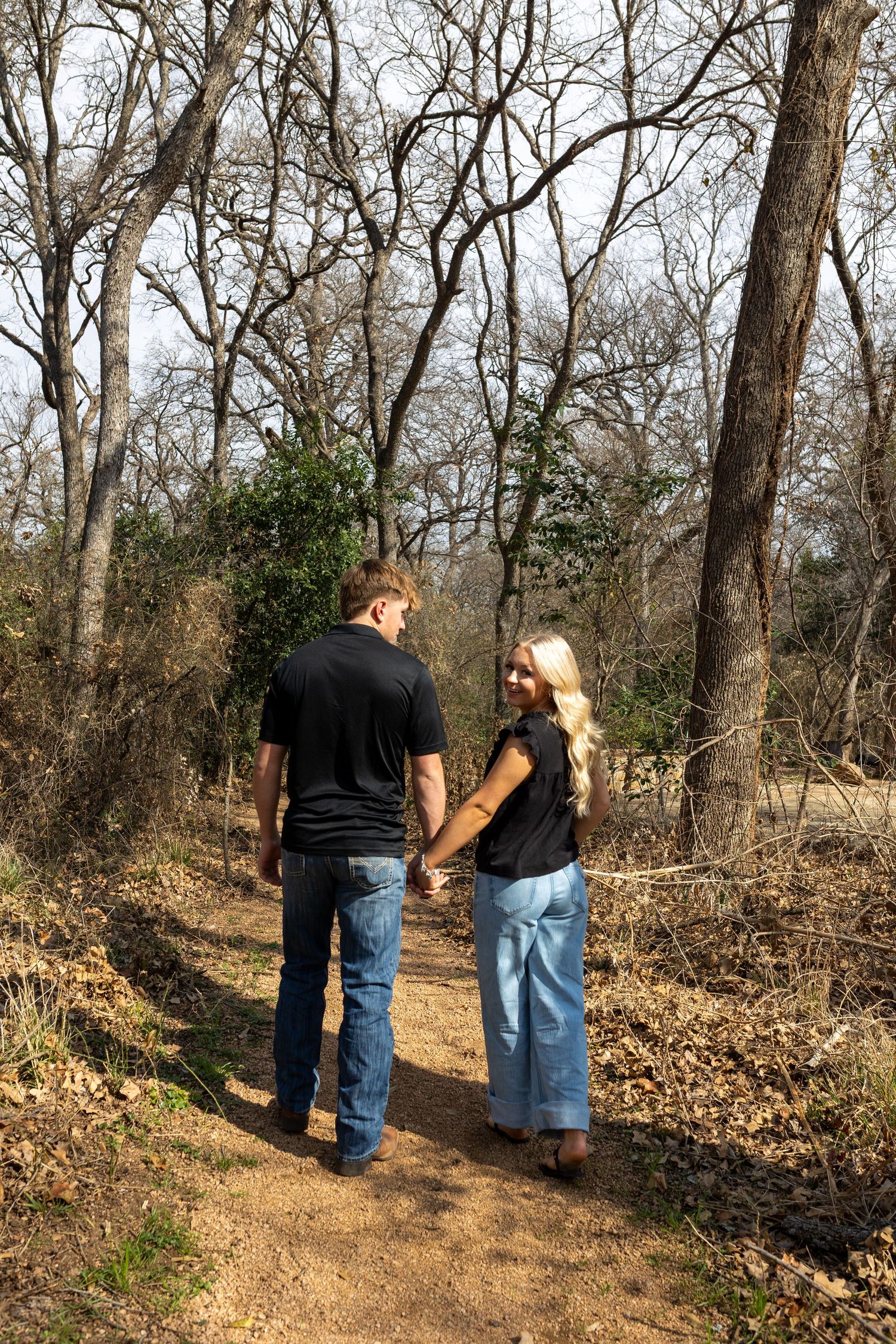 A couple walks hand in hand along a wooded trail during winter with bare trees in the background.
