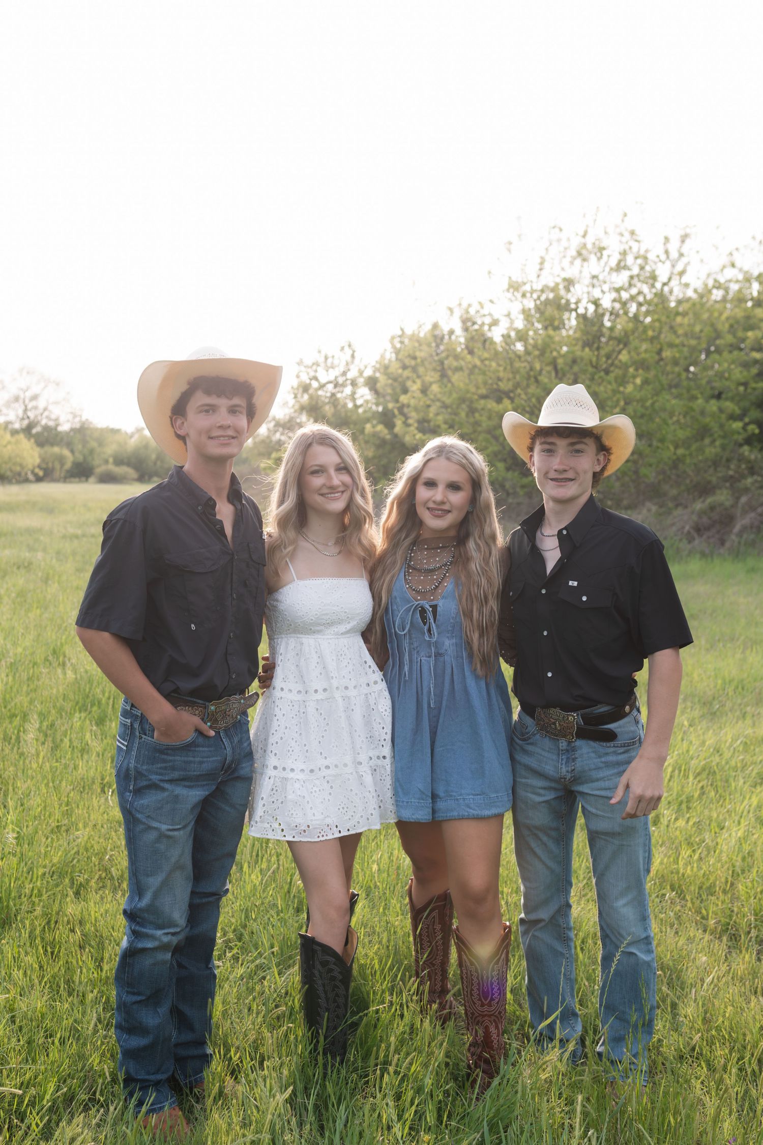Four young people in cowboy hats and boots pose in a green field during golden hour.