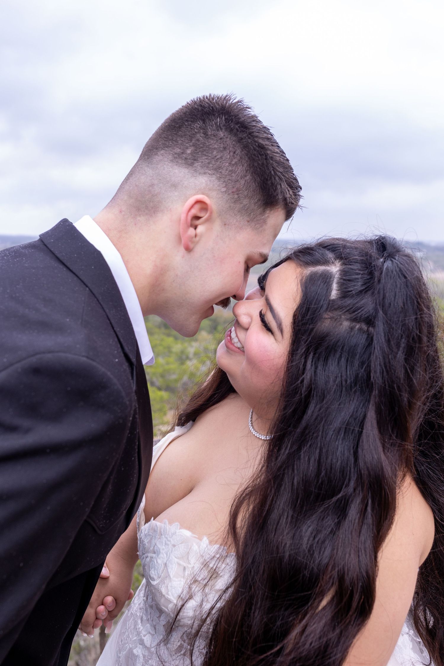 A bride and groom share a romantic nose-to-nose moment outdoors, dressed in wedding attire against a cloudy sky.