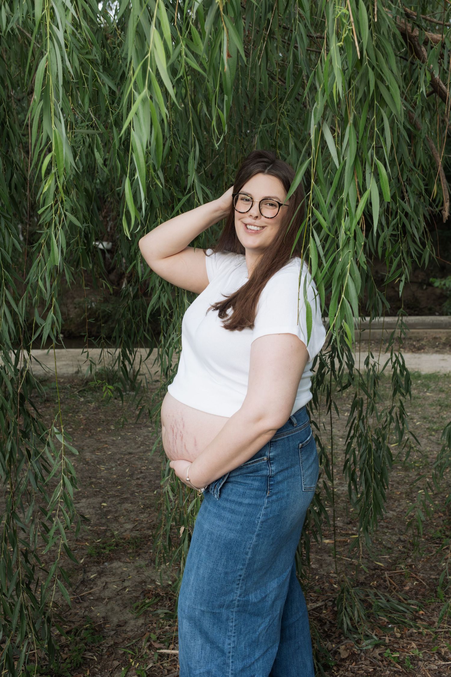 Smiling pregnant woman in white tee and jeans poses outdoors under weeping willow tree, showing bare baby bump.