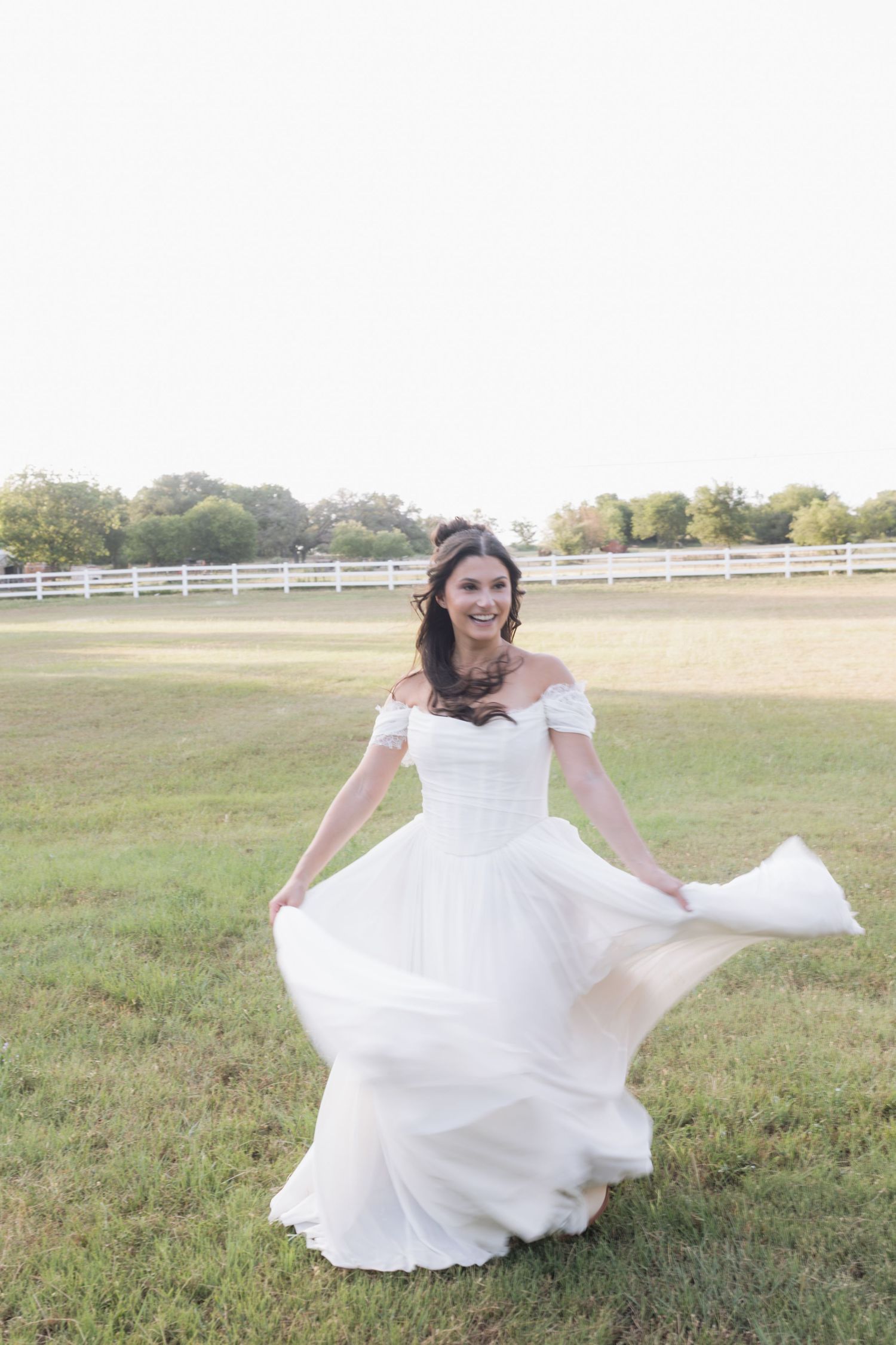 Smiling bride in off-shoulder white gown twirling dress in open field at golden hour wedding.