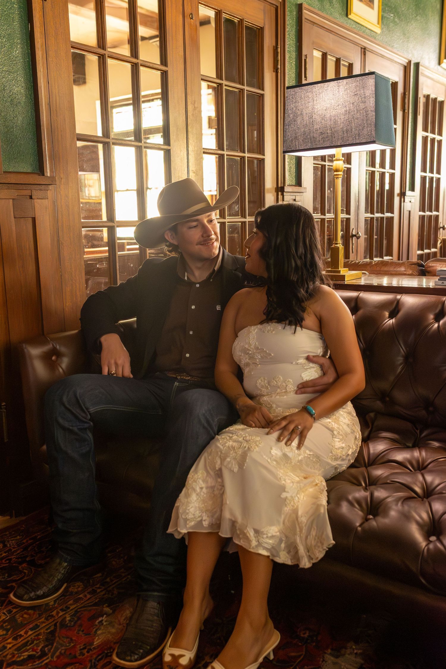 A couple sits together on a brown leather couch in a warmly lit room with wooden paneled walls.