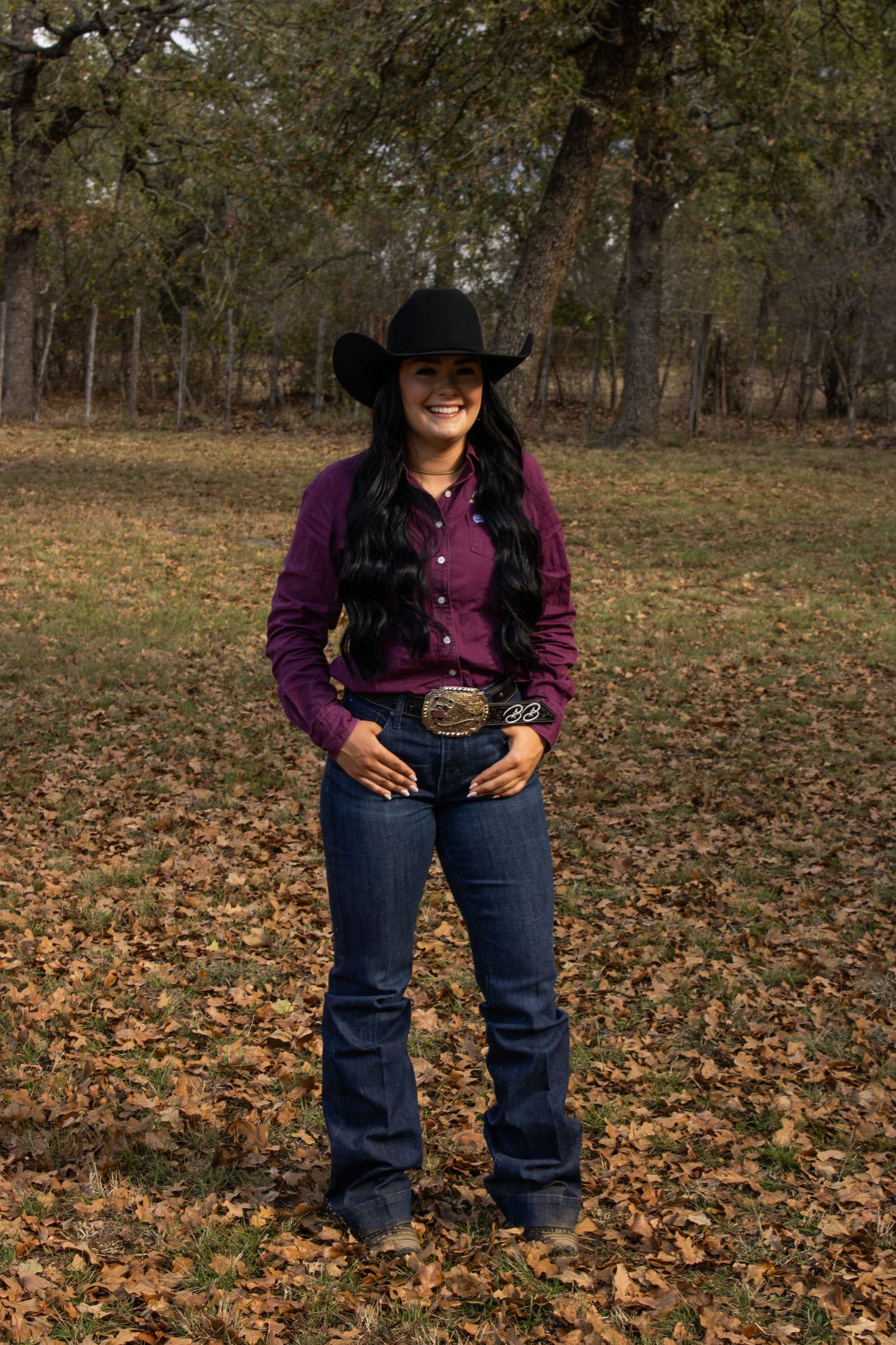 A country western ensemble with dark denim jeans, decorative belt buckle, and purple shirt against autumn foliage.