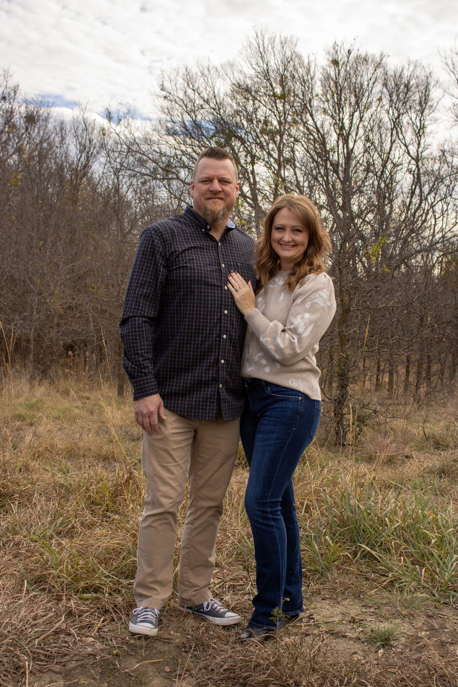 A couple stands together in a natural outdoor setting with bare trees and tall grass in the background.