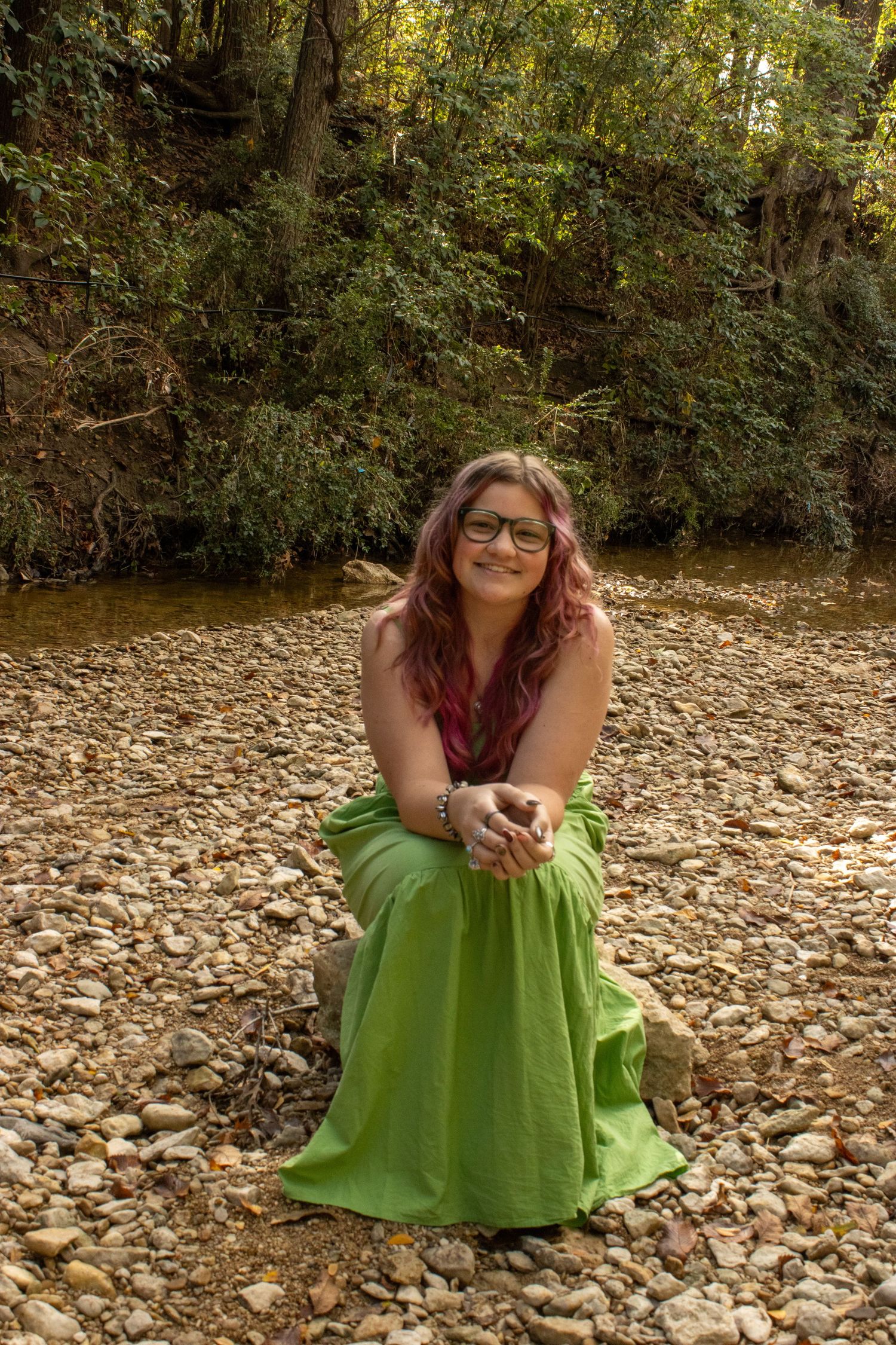 A person in a bright green dress sits on a rocky riverbed surrounded by autumn leaves and forest scenery.