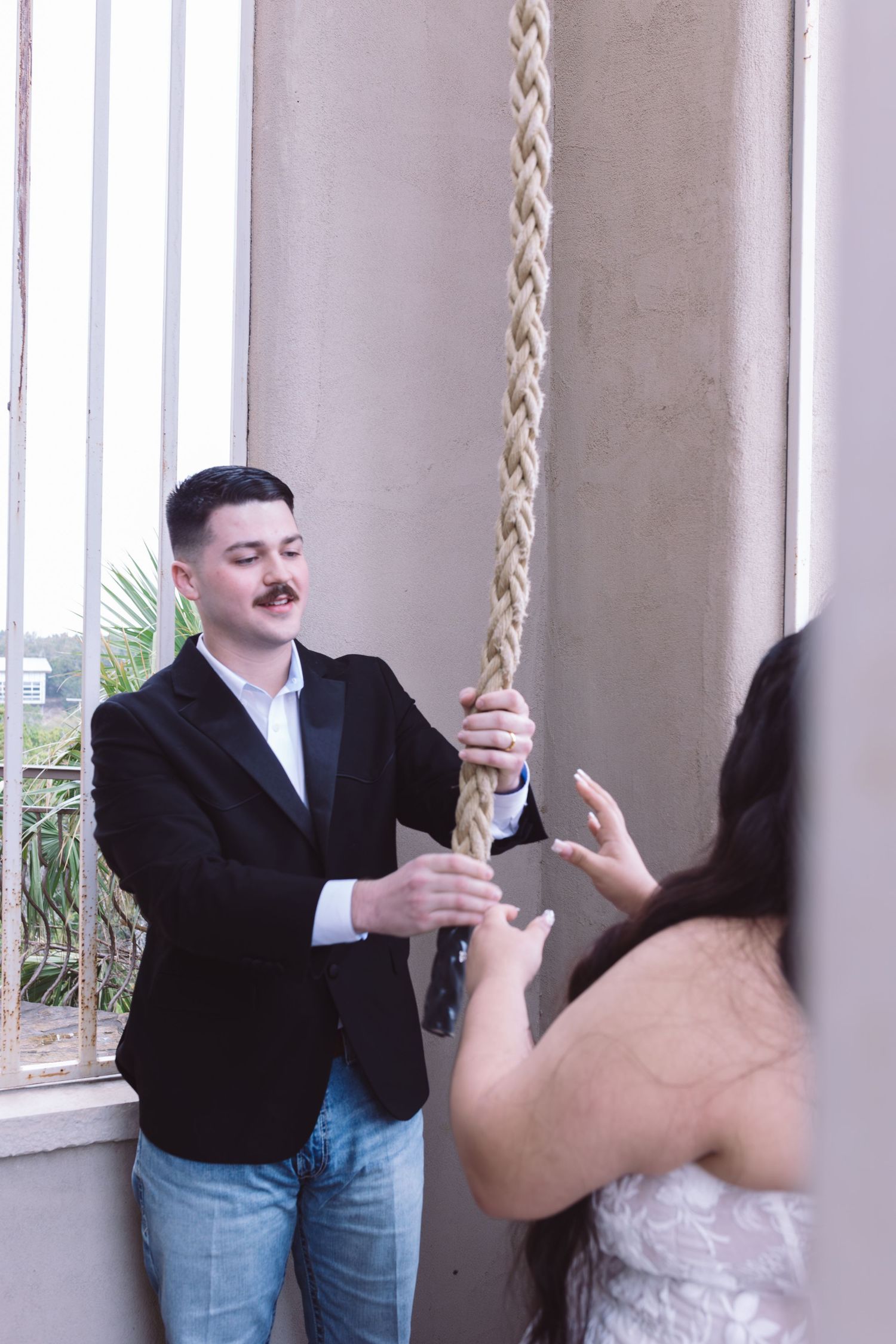 A man in a black blazer and jeans holds a braided rope while a woman in a white dress reaches toward it indoors.