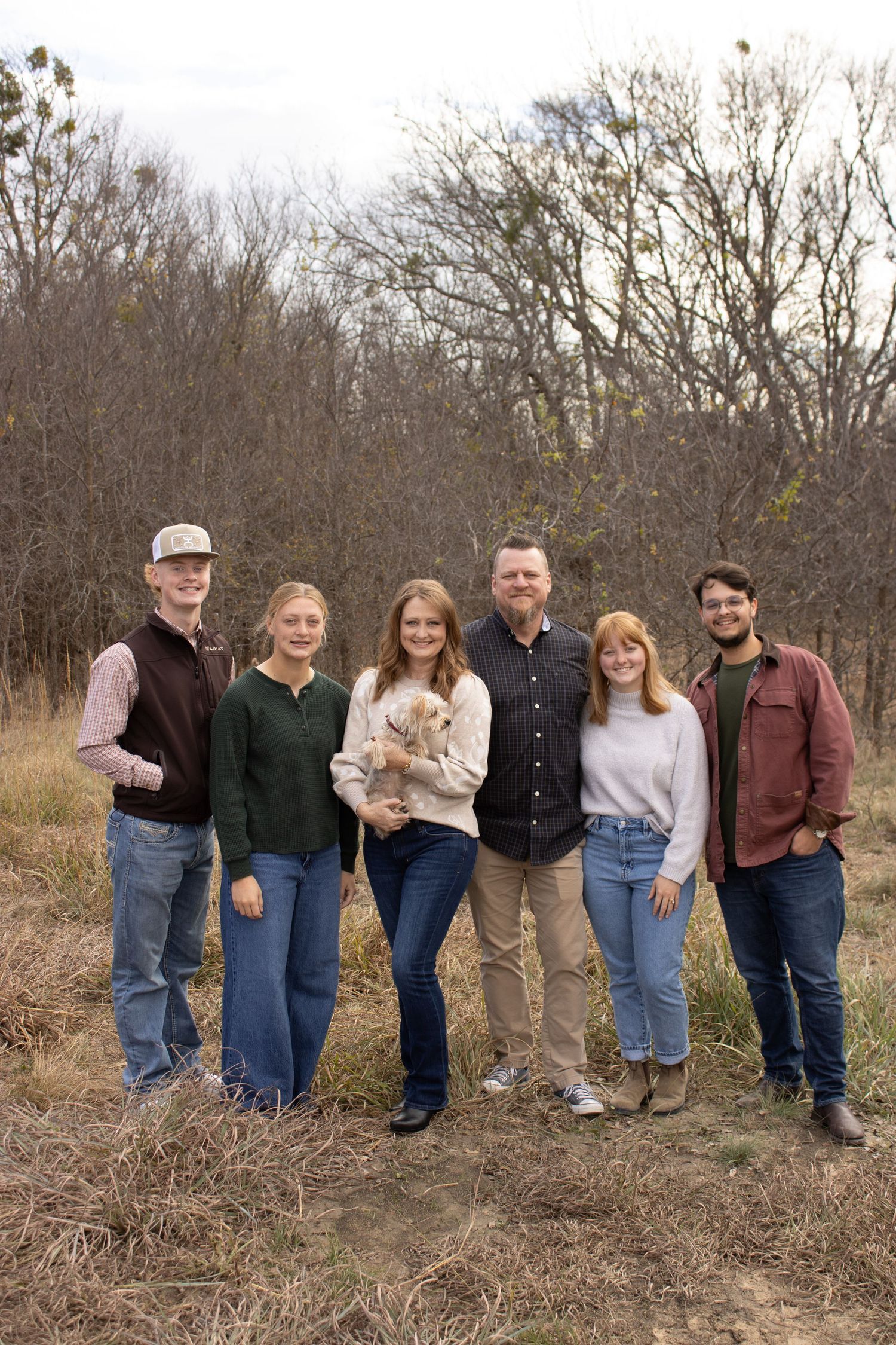 A group of people standing together in casual attire against a backdrop of bare autumn trees and brush.
