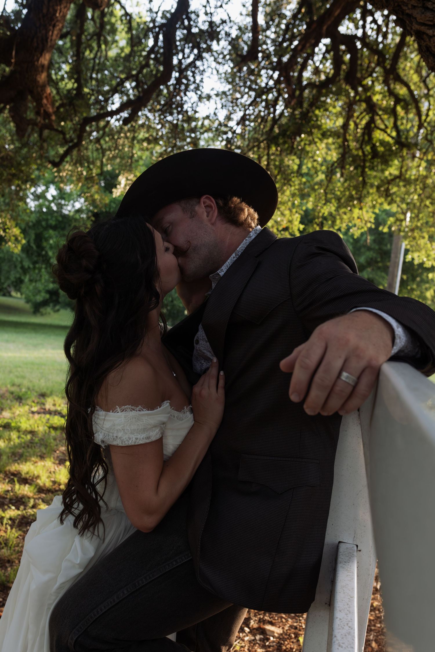 Newlyweds sharing a kiss under a tree, groom in dark suit and cowboy hat, bride in white off-shoulder gown.
