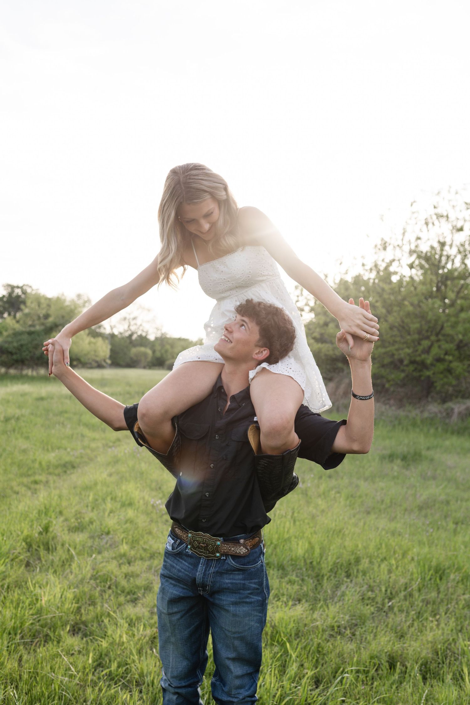 Young couple in a sunny green field, man lifting woman on his shoulders while holding hands outdoors.
