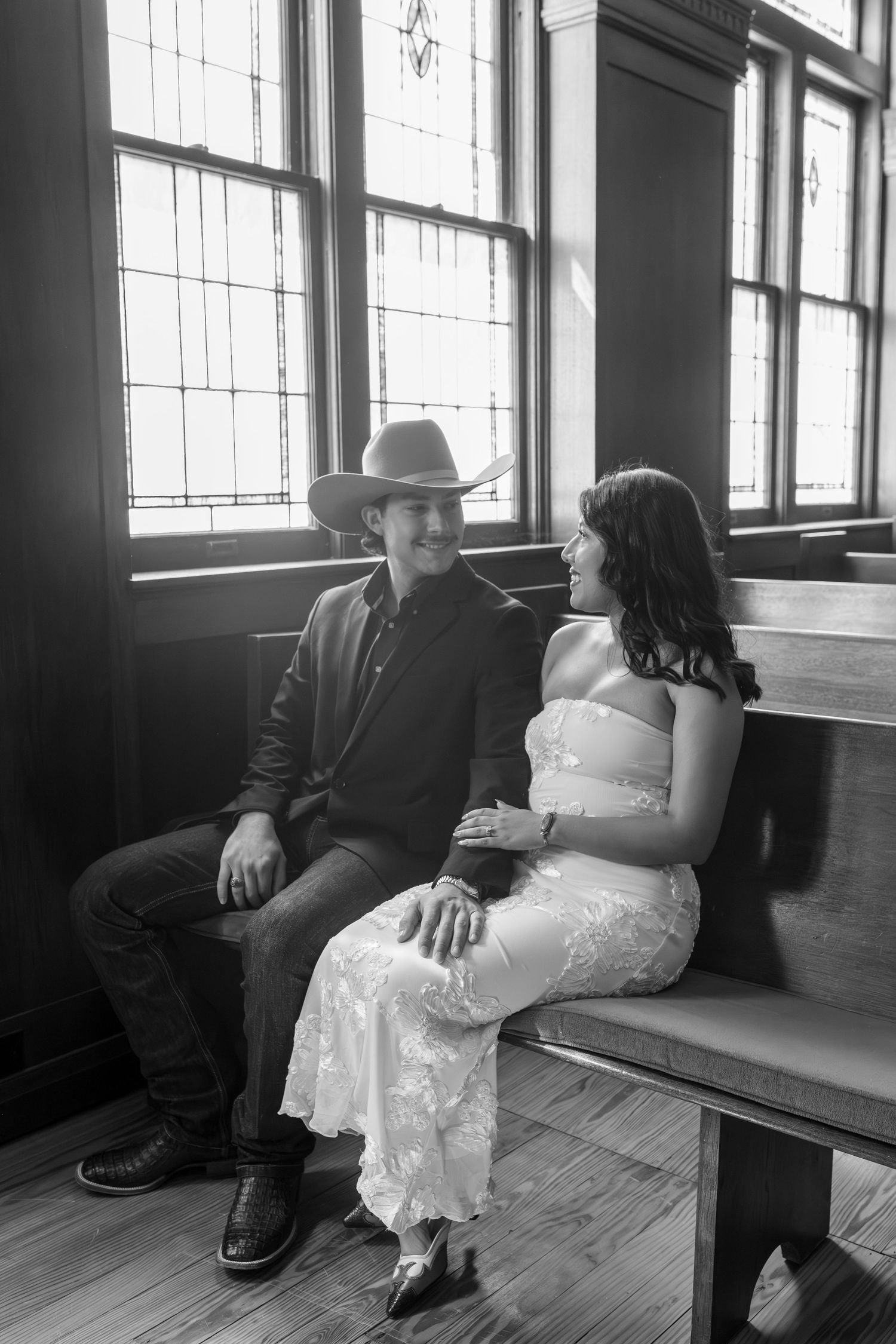 Black and white photo of a couple in wedding attire sitting on a church pew, man in cowboy hat, woman in white lace dress.