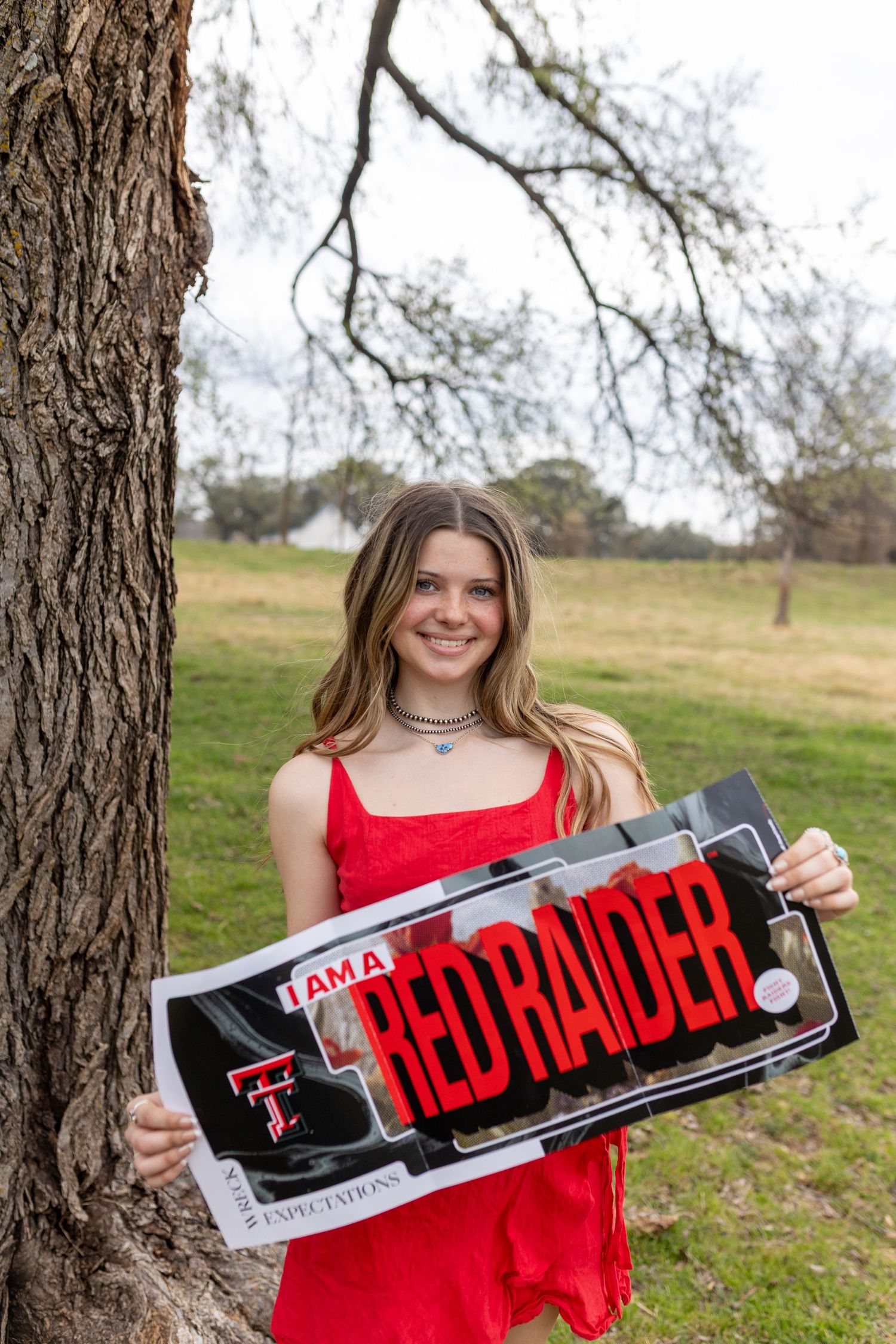 Someone in a red dress holds up a skateboard deck with red and black graphics against a natural outdoor backdrop.