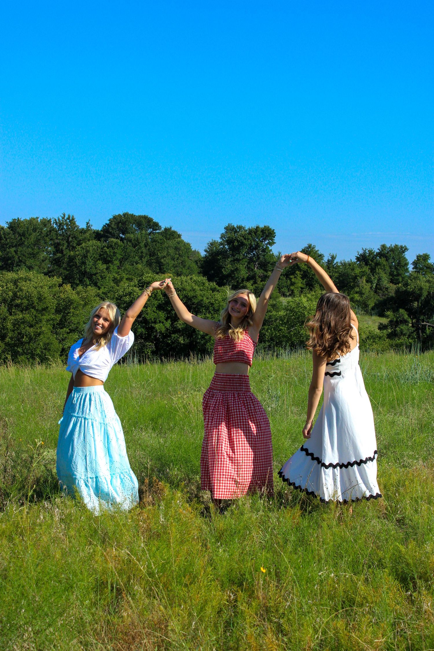 Group in summer dresses posing with raised arms in green field under blue sky.
