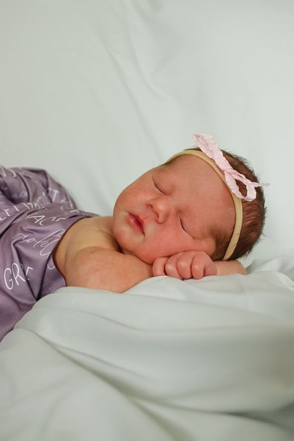 Peaceful newborn baby sleeping on white sheets wearing purple outfit and pink bow.