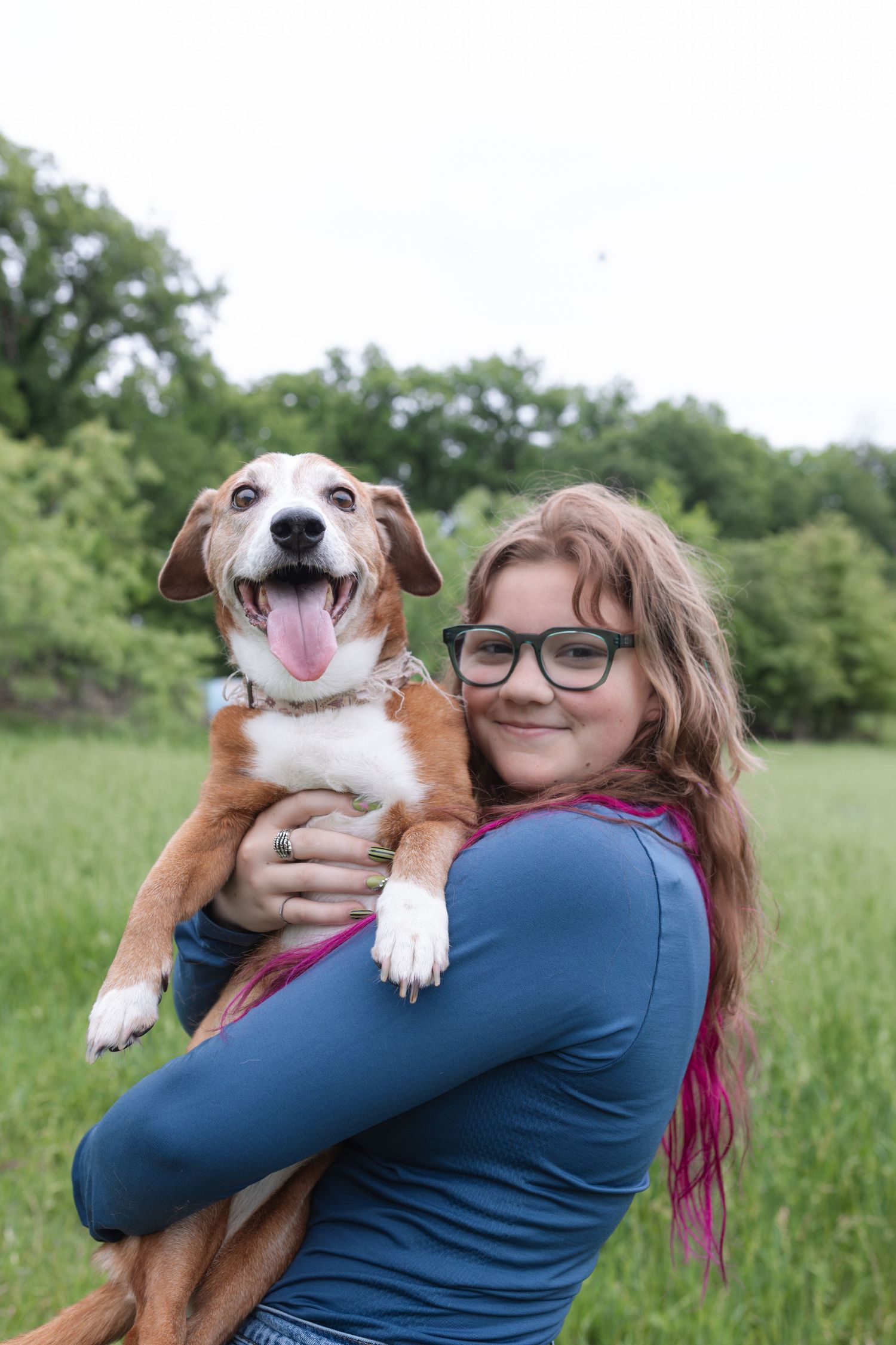 Smiling girl with glasses holds a happy brown and white dog with tongue out in a green outdoor field.