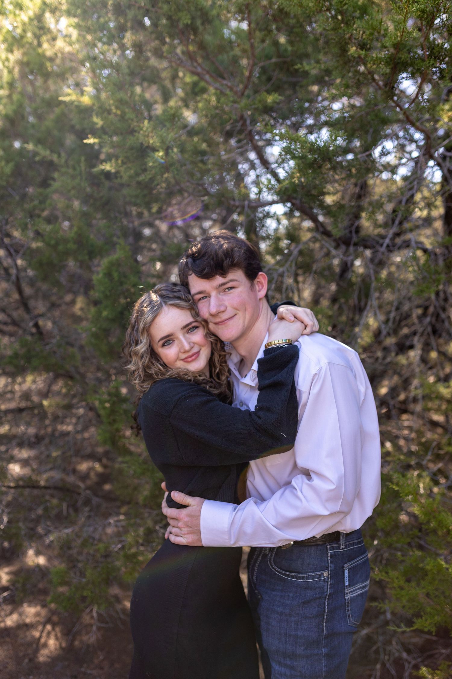 A couple embraces outdoors in a natural setting with trees and foliage in the background.