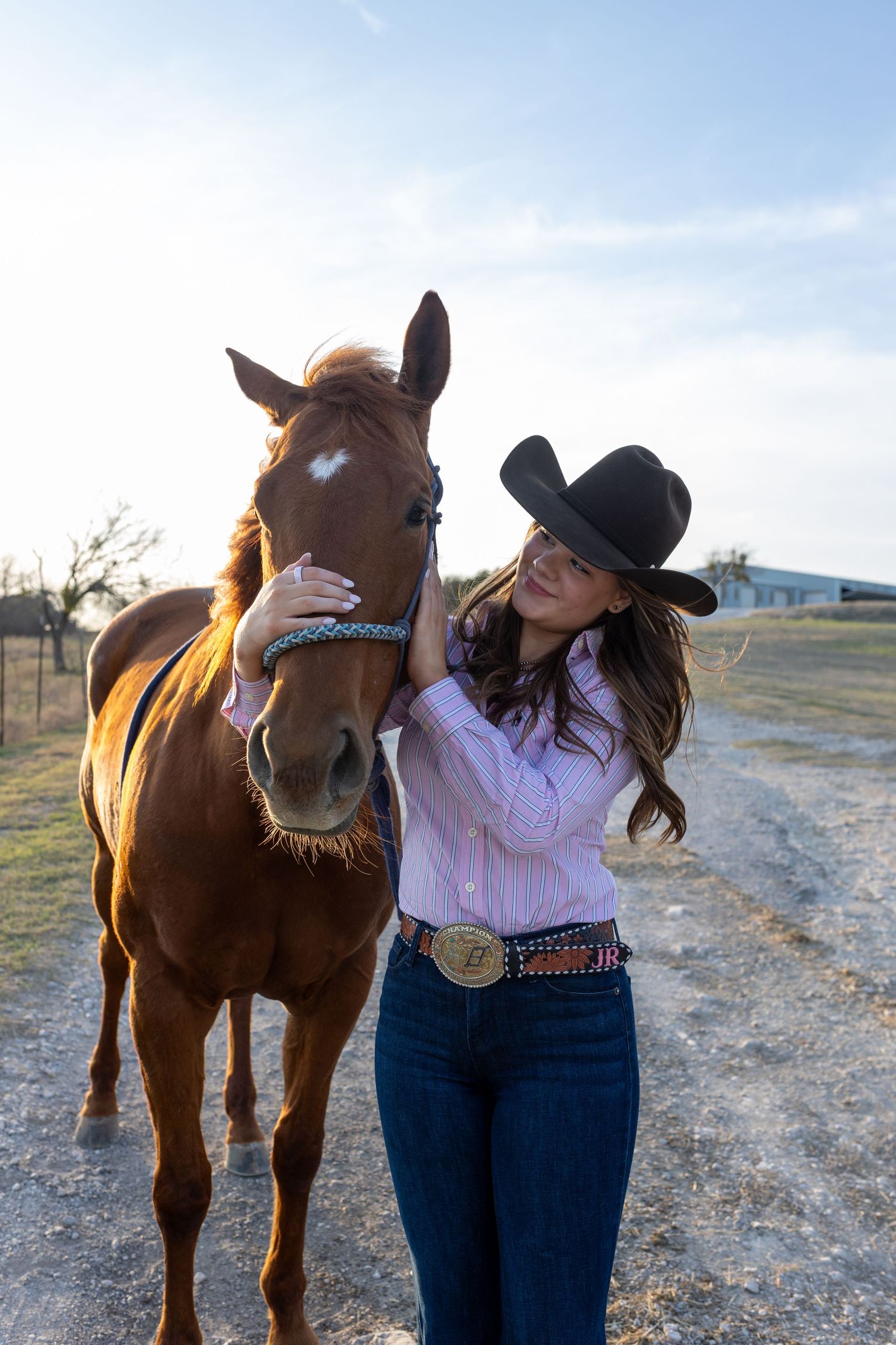 A horse and rider in western attire share a tender moment in an open field during golden hour lighting.