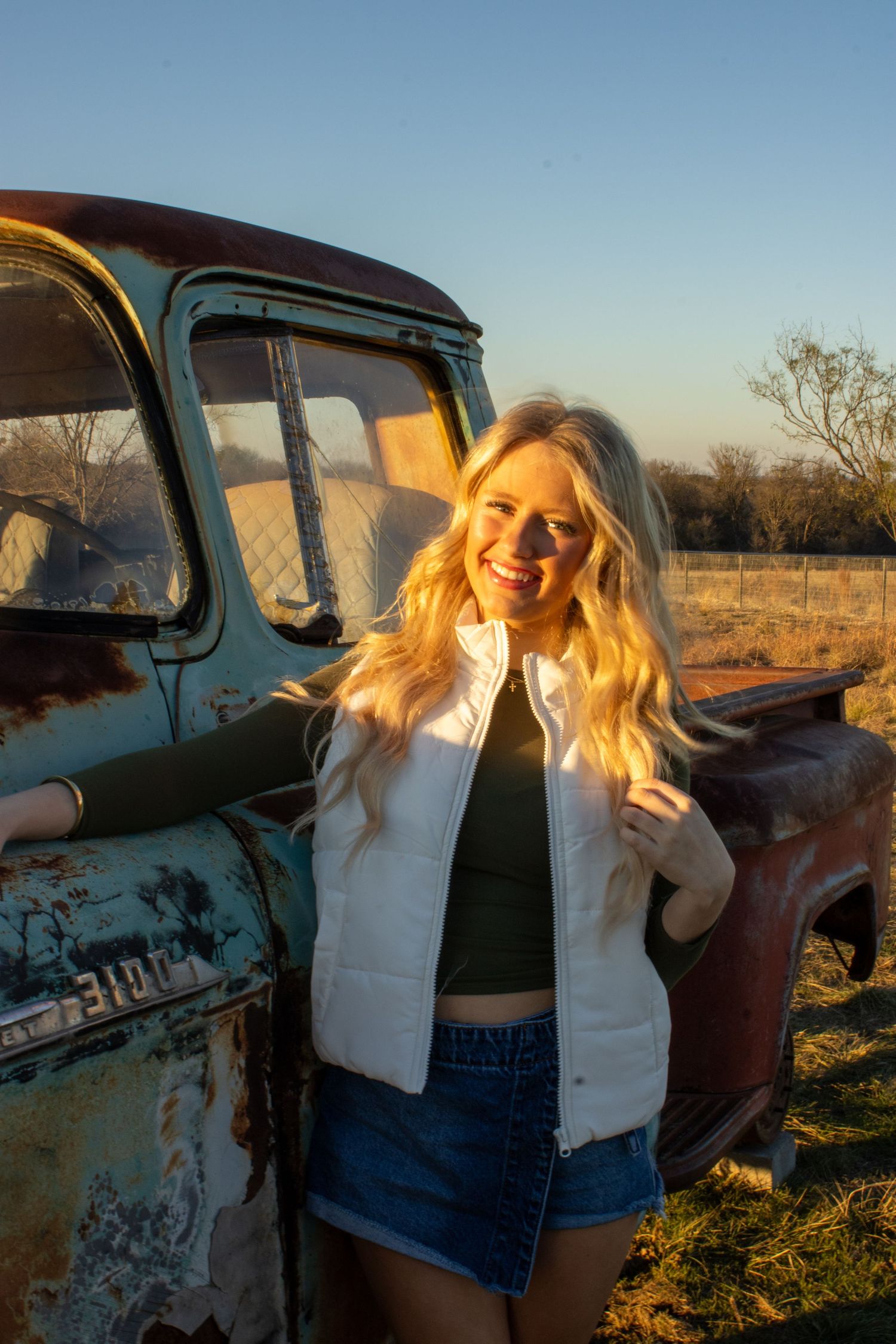 Two people laugh and pose together near a vintage rustic truck at golden hour in a rural setting.