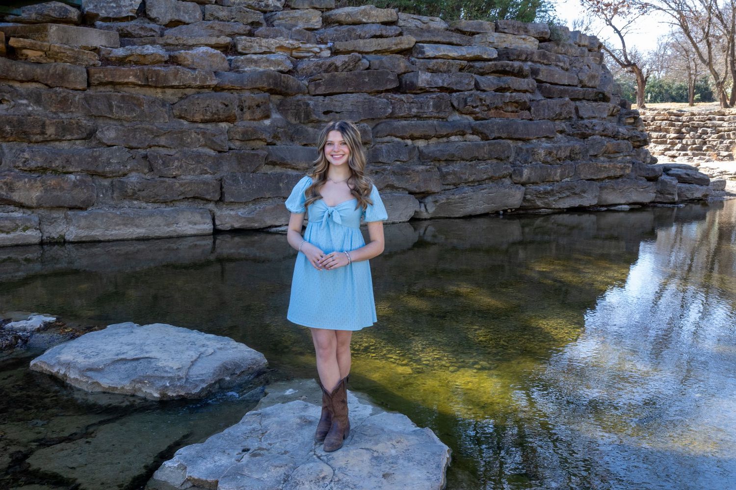 Stone retaining wall reflects in calm pond water with natural landscaping and greenery in background.