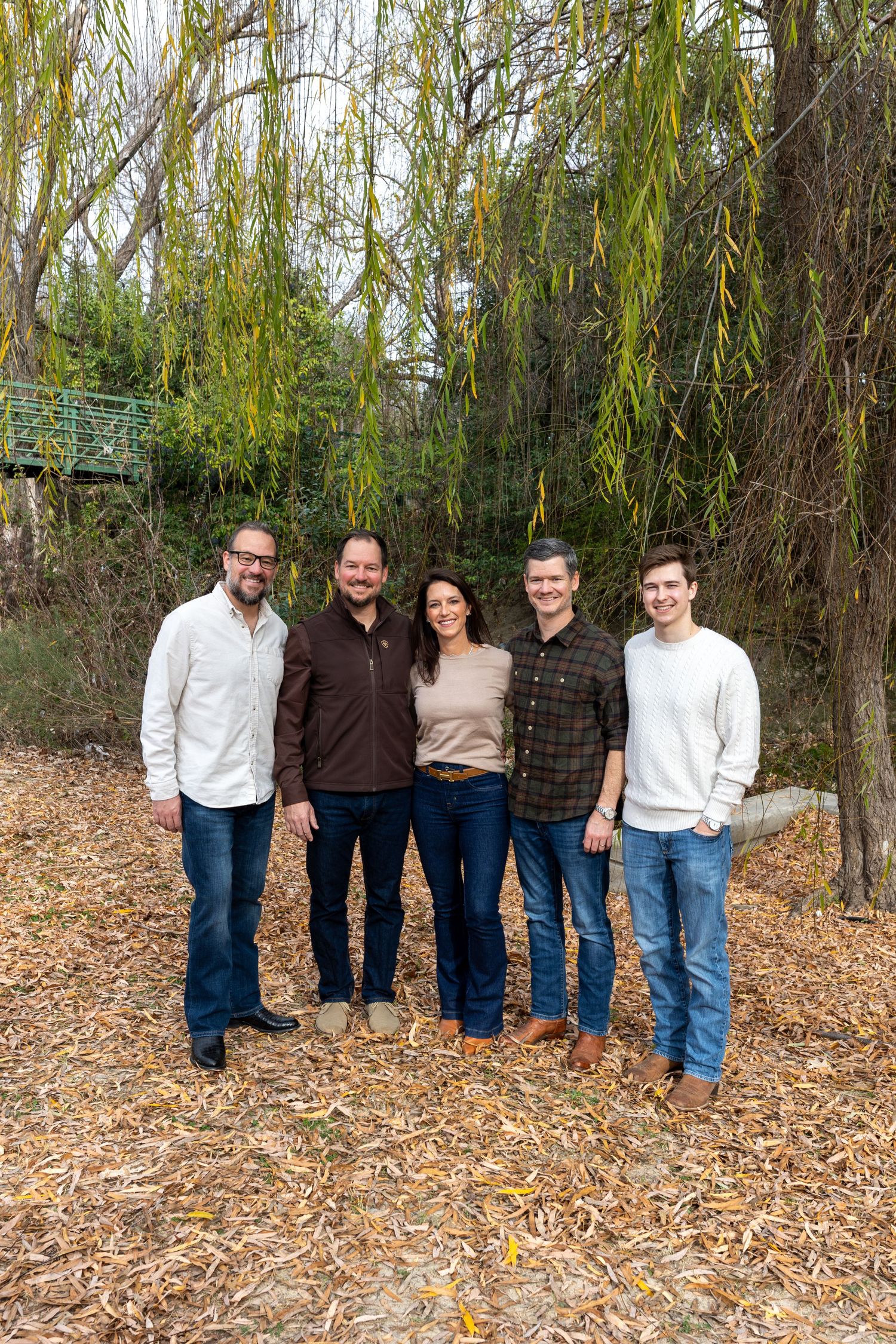 A group of people standing together outdoors under willow trees on a bed of fallen autumn leaves.