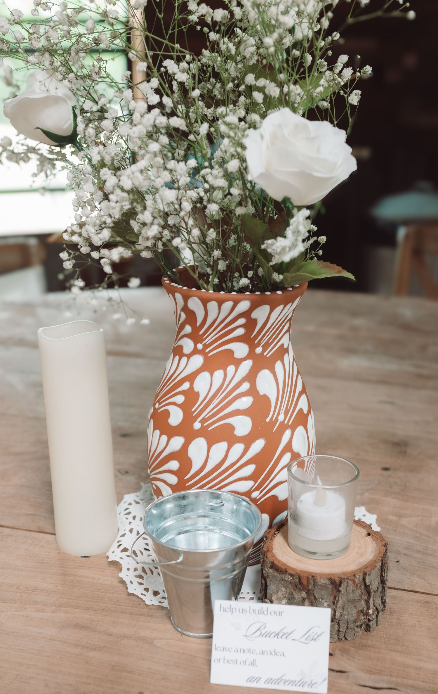Rustic wedding centerpiece with terracotta floral vase, white roses, baby's breath, candles, and a small bucket on wood table.