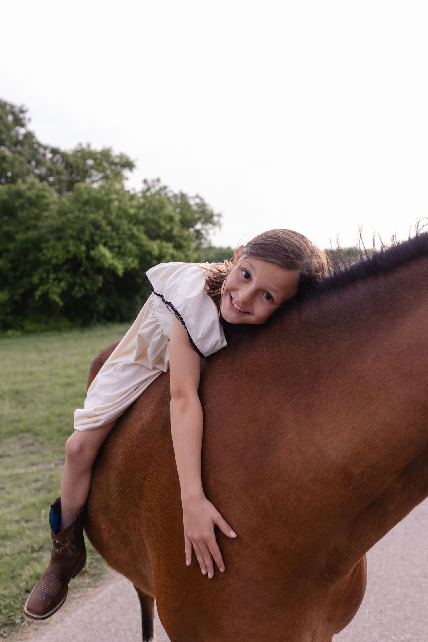 Young girl in white dress hugging a brown horse outdoors, smiling warmly in a green countryside setting.