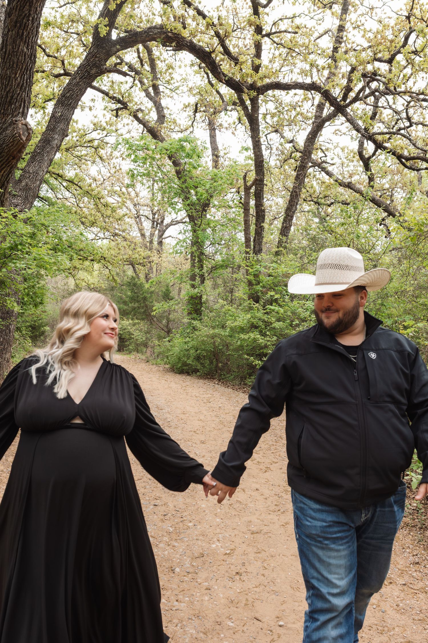 A couple in dark clothing holds hands and laughs together on a leaf-covered forest path.